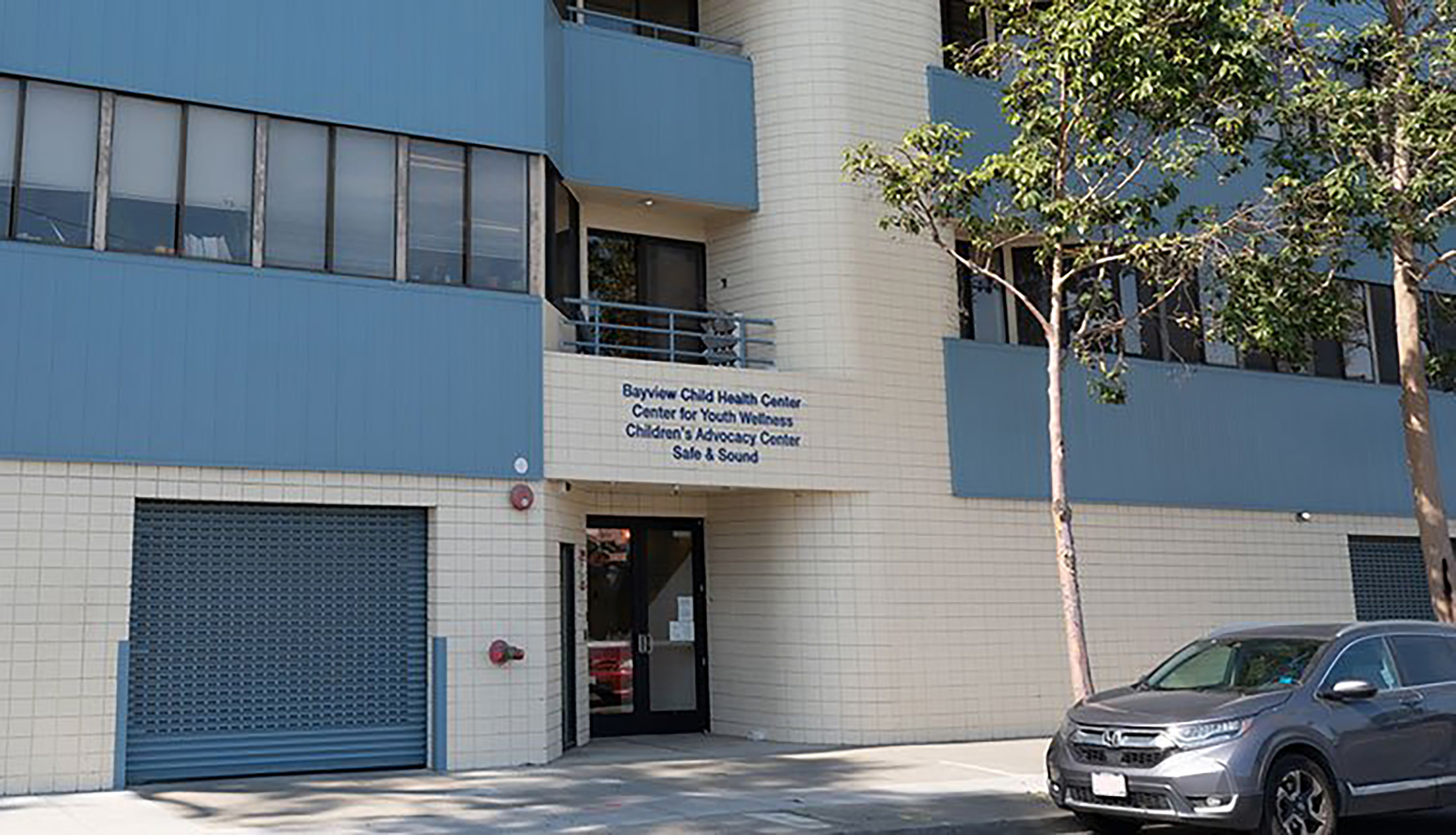 Exterior view of the Bayview Child Health Center building entrance, with signage listing children’s health and advocacy services, blue facade panels, and a parked car along the street.