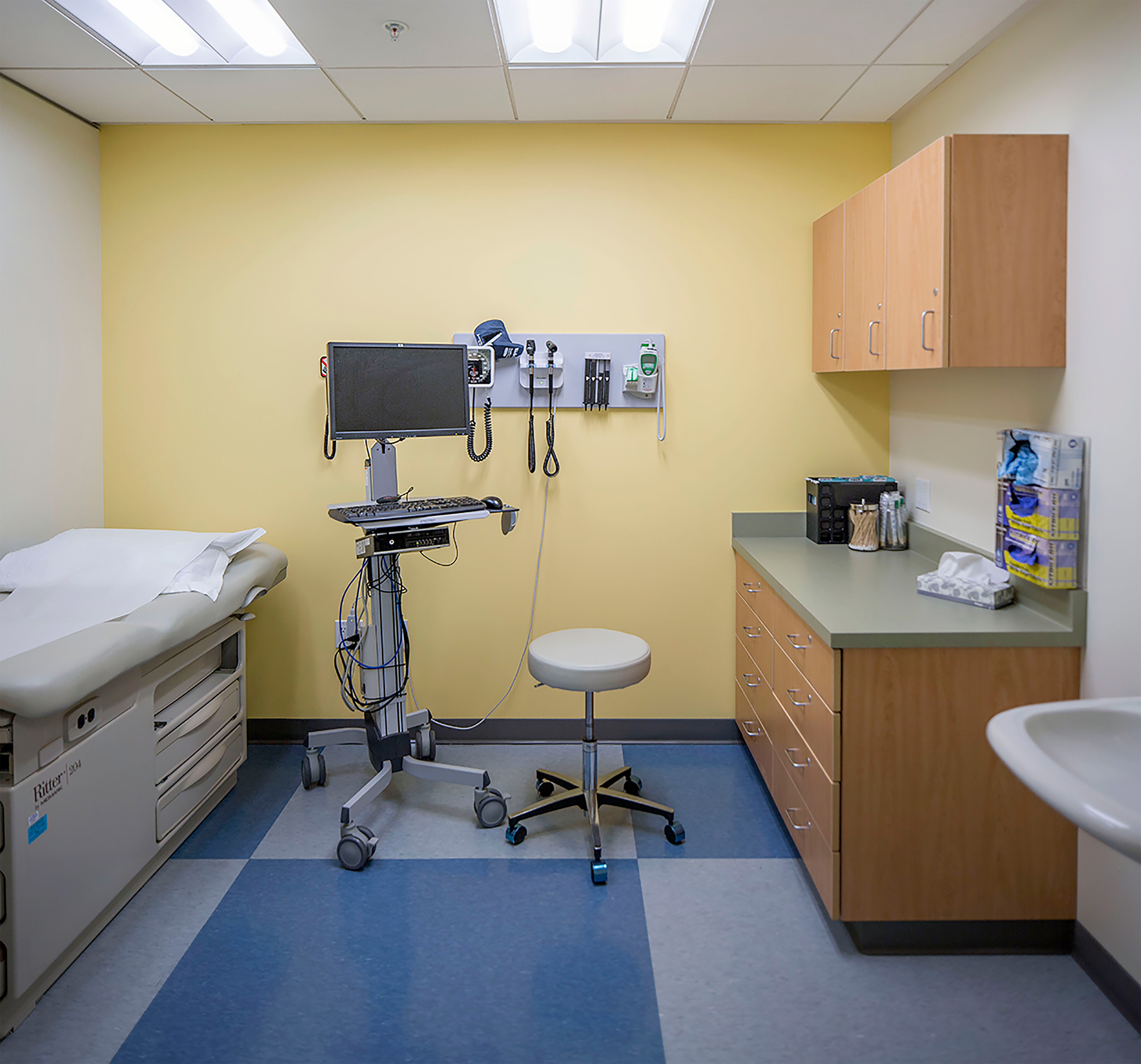 Pediatric exam room with an exam table, medical equipment, rolling stool, sink, and wood storage cabinets against a yellow wall.
