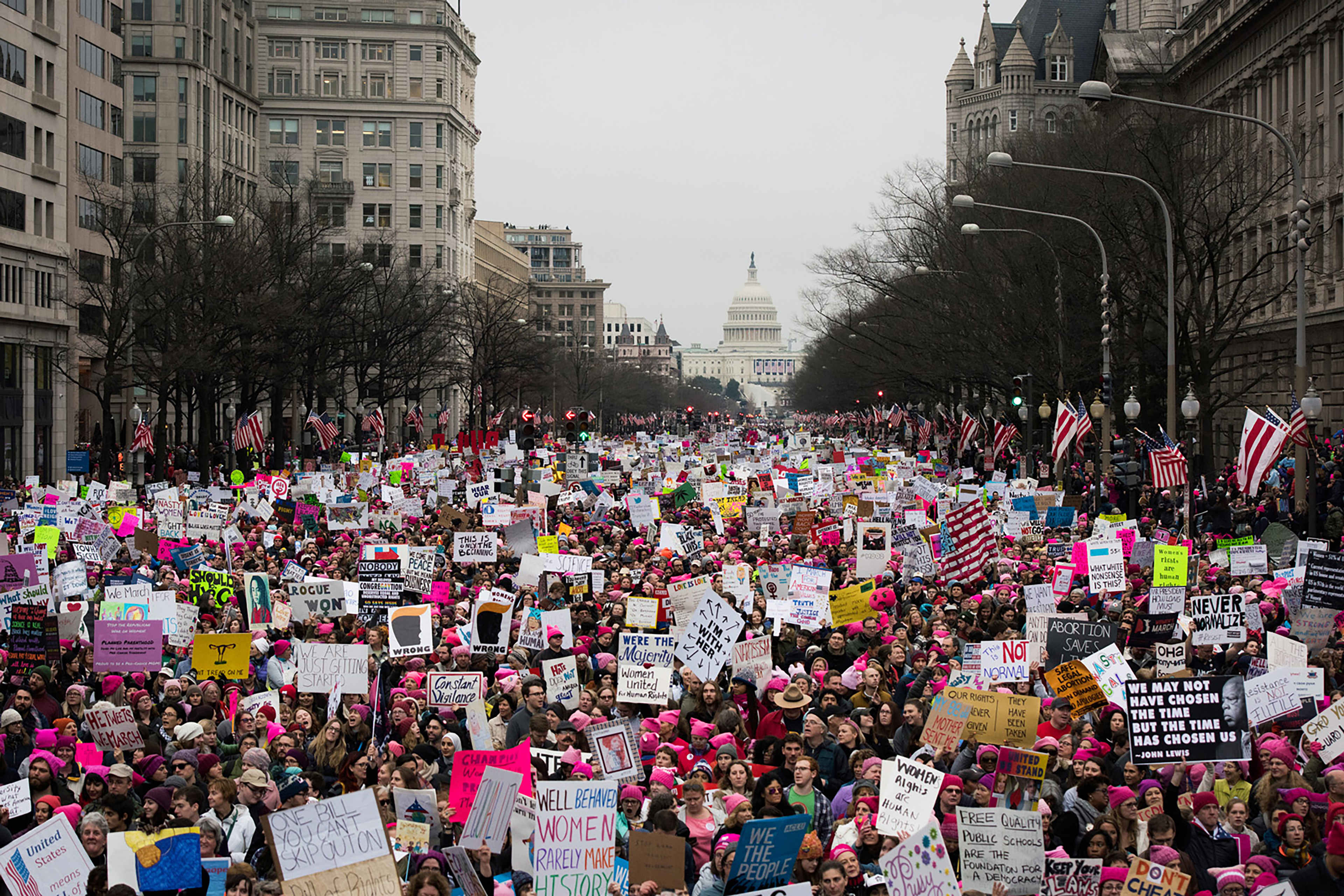 A large crowd fills a broad Washington, D.C., avenue during the Women’s March, holding colorful protest signs and American flags. Leafless trees and historic buildings frame the street, with the U.S. Capitol visible in the distance.