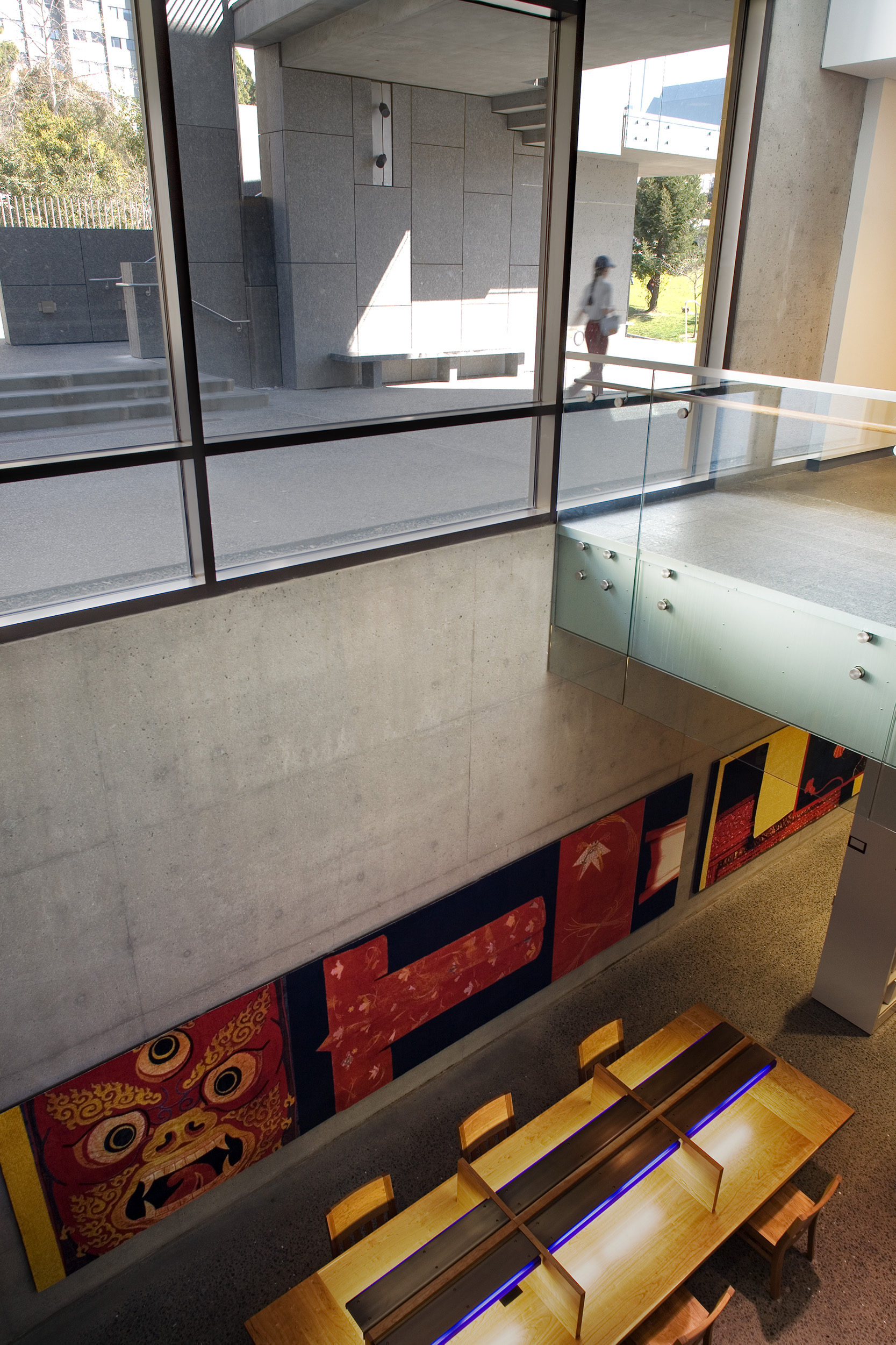Double-height interior with exposed concrete walls, a glass-railed bridge, and large windows overlooking an outdoor courtyard. A long textile artwork and illuminated communal table occupy the lower level, while a motion-blurred person passes outside.