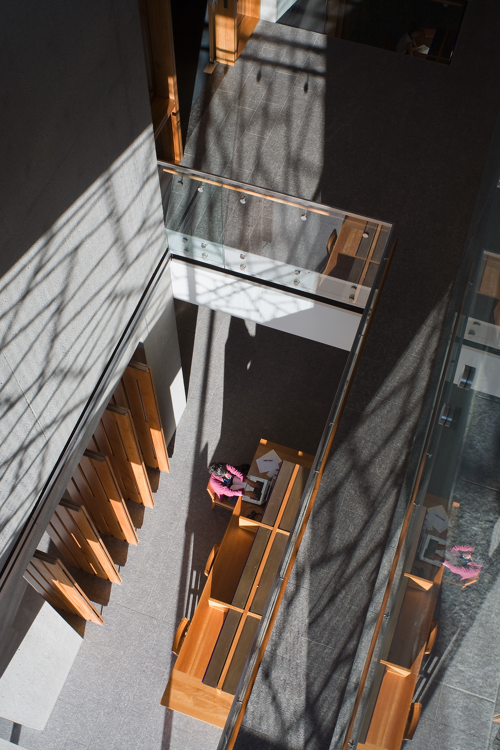 Overhead view into a multilevel concrete interior with glass railings and long wood study tables. A person works alone at one table while strong daylight creates dramatic geometric shadows across the walls and floors.