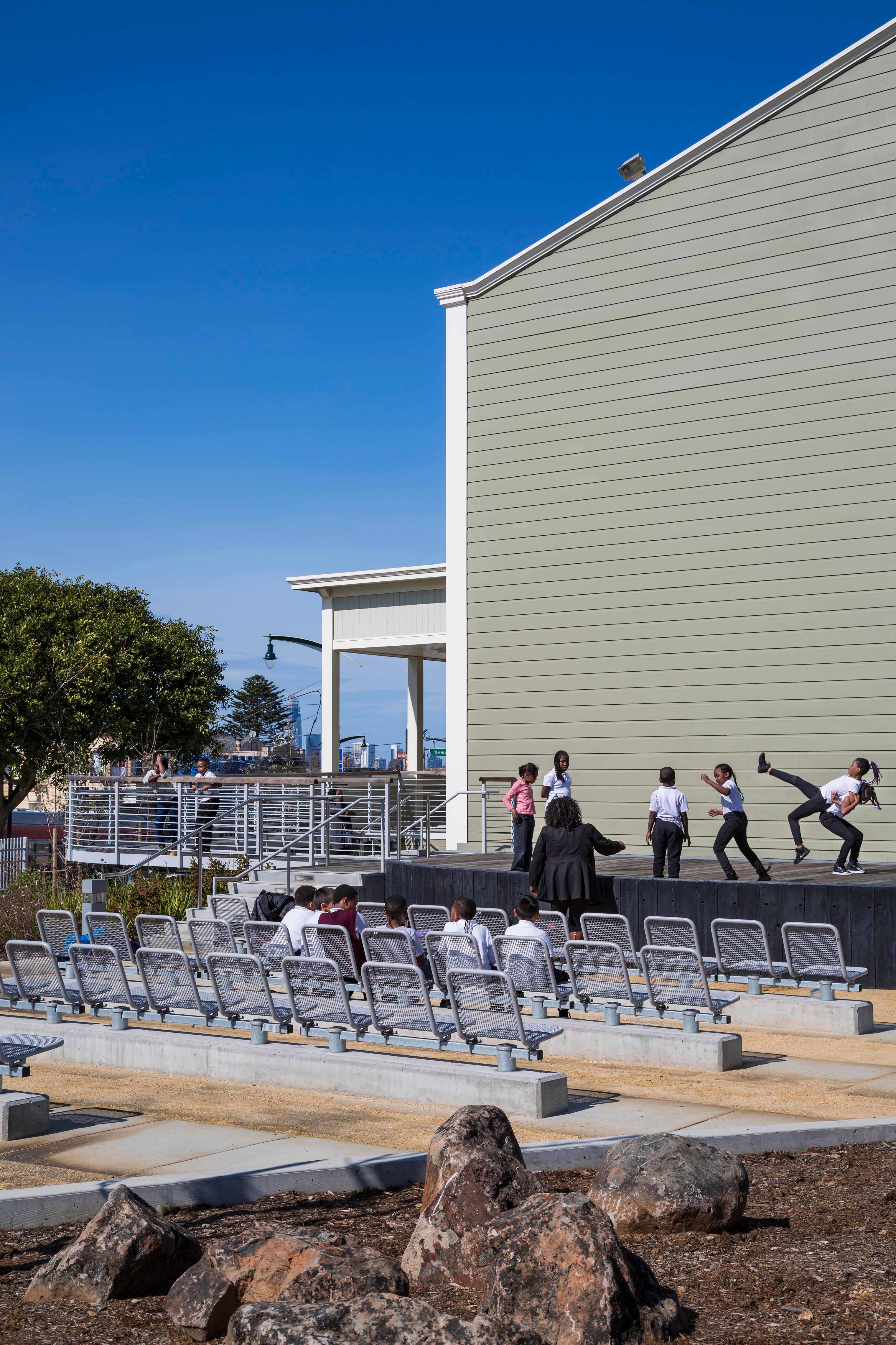 Outdoor amphitheater beside the San Francisco Opera House, with children performing on a raised stage while an audience sits in metal seating under a clear blue sky.