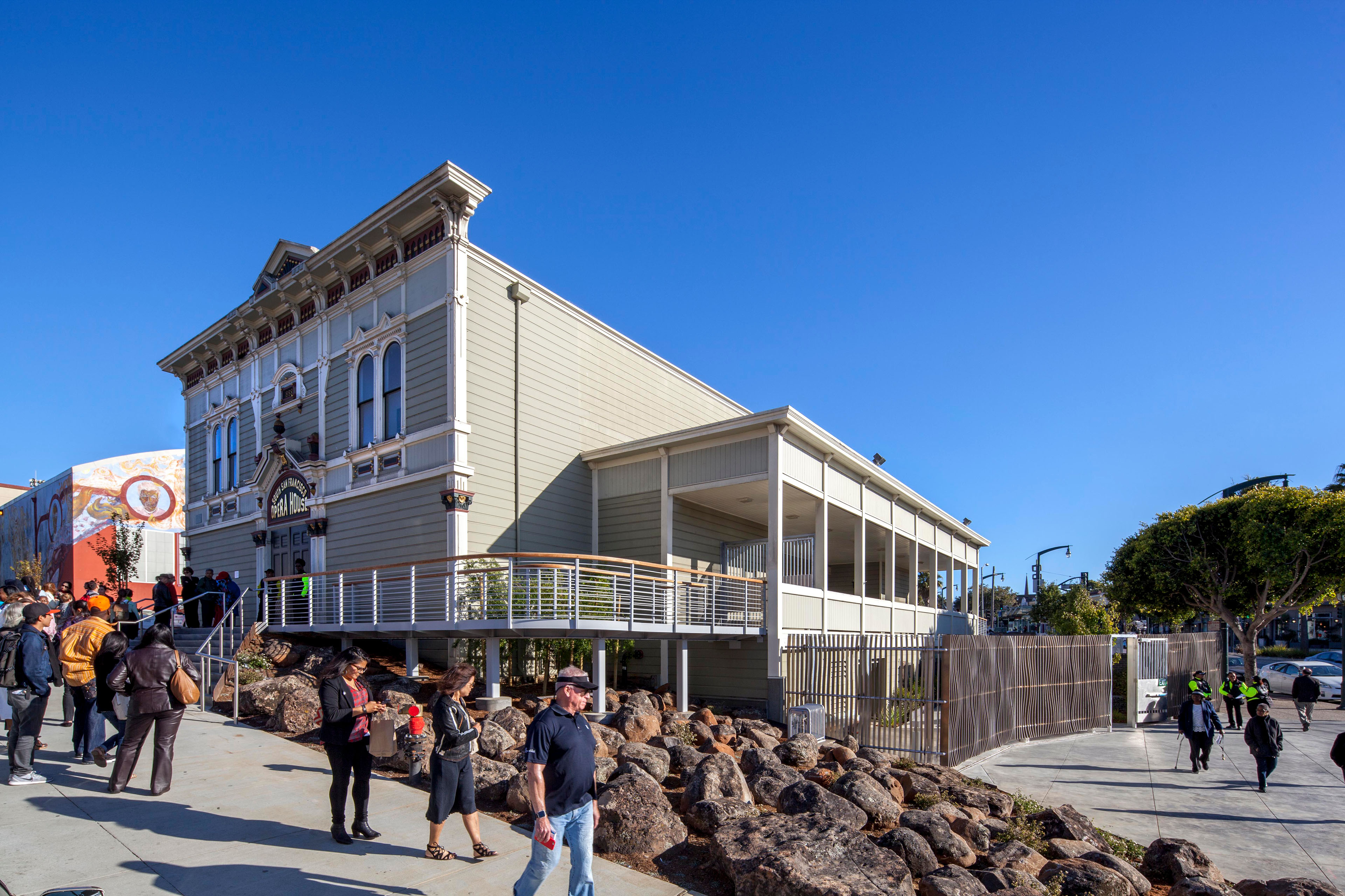 Exterior view of the San Francisco Opera House on a sunny day, showing the restored historic facade, adjacent covered porch, accessible ramp, landscaped rock slope, and pedestrians gathered along the sidewalk.