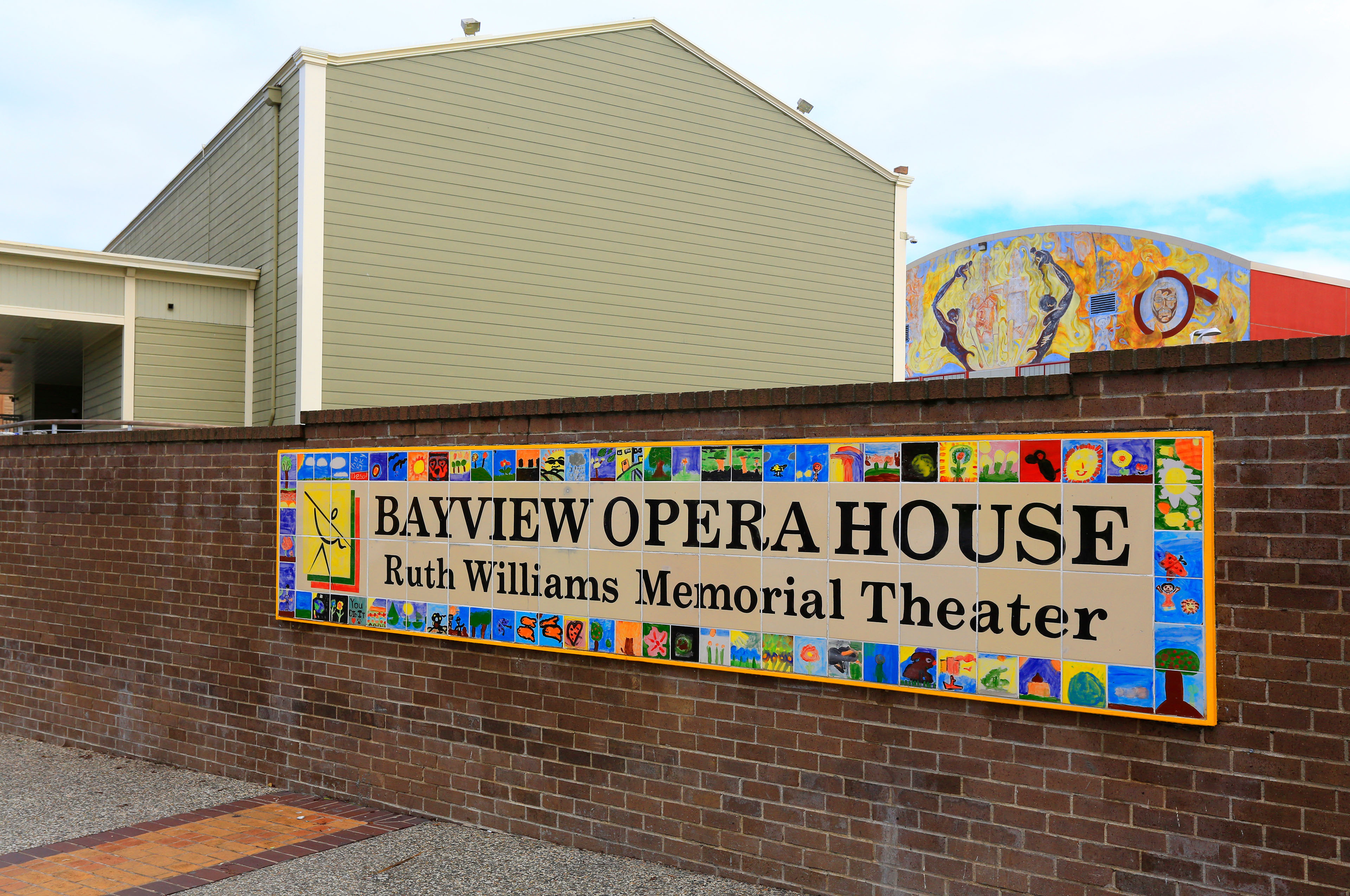 Colorful tile sign on a brick wall reading “Bayview Opera House Ruth Williams Memorial Theater,” with the building and a painted mural visible in the background.