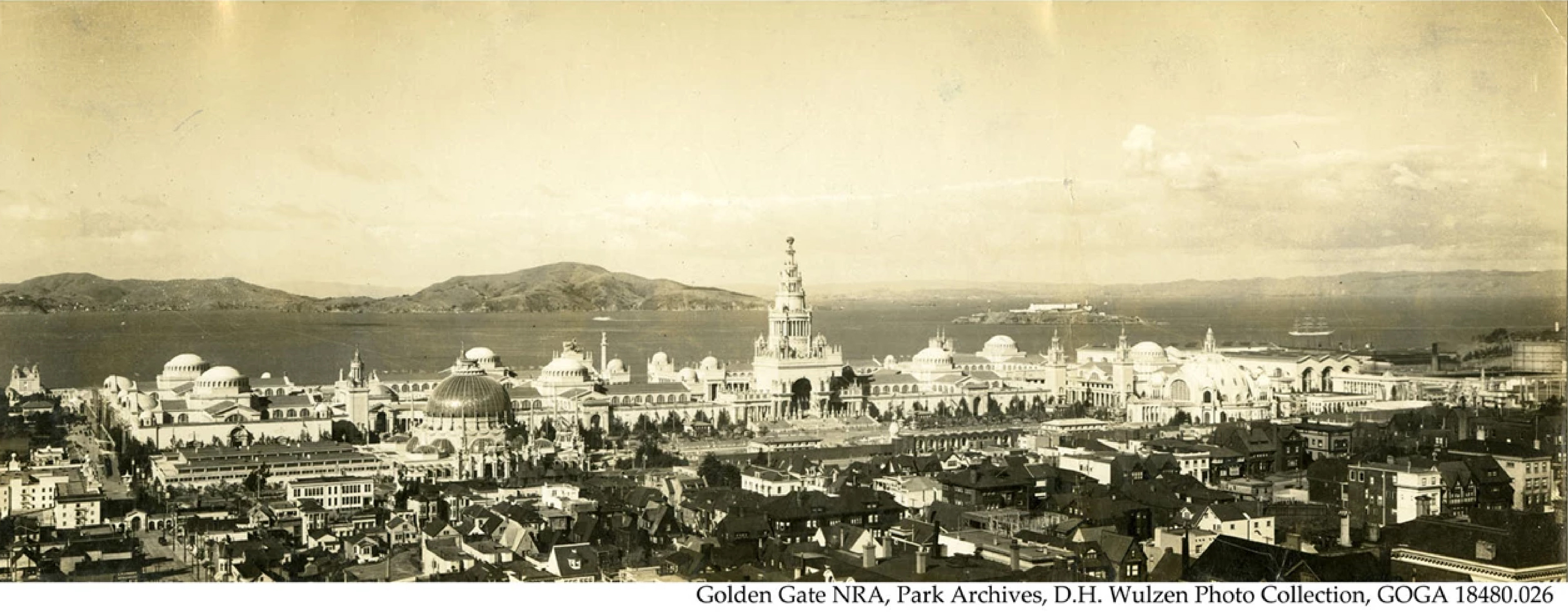 Sepia-toned panoramic photograph of the 1915 Panama-Pacific International Exposition in San Francisco. Large domed exhibition halls and a tall central tower stretch across the middle ground, with dense residential buildings in the foreground and the bay, islands, and distant hills beyond.