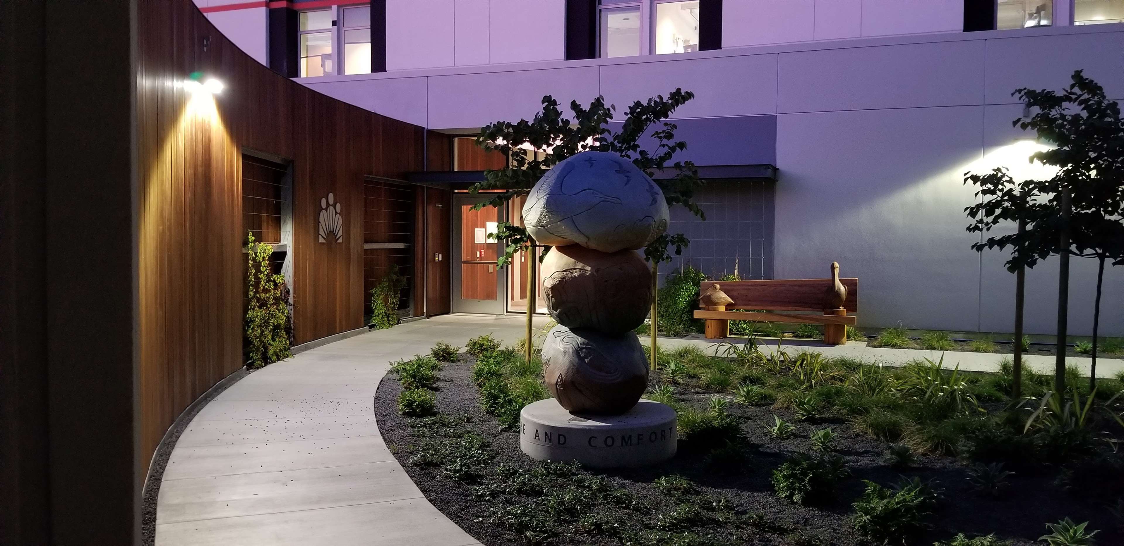 Nighttime view of a behavioral health healing garden with a curving walkway, illuminated wood-clad walls, landscaped planting, carved benches, and a stacked wildlife sculpture.