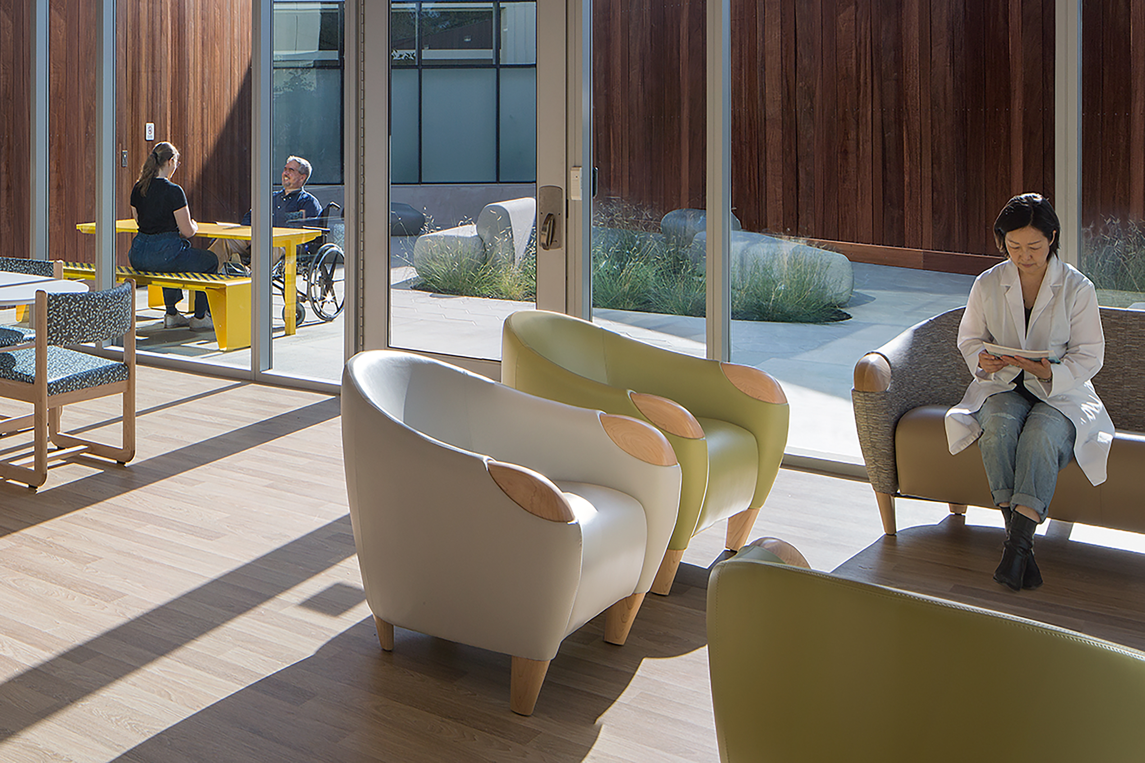 alt text Sunlit behavioral health lounge with upholstered seating, floor-to-ceiling glass doors opening to a landscaped courtyard, a clinician using a tablet, and two people seated outside.