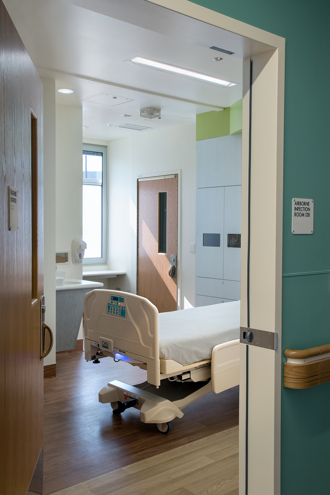 Airborne infection patient room with a hospital bed, built-in medical services, wood-look flooring, and natural light from a tall window.