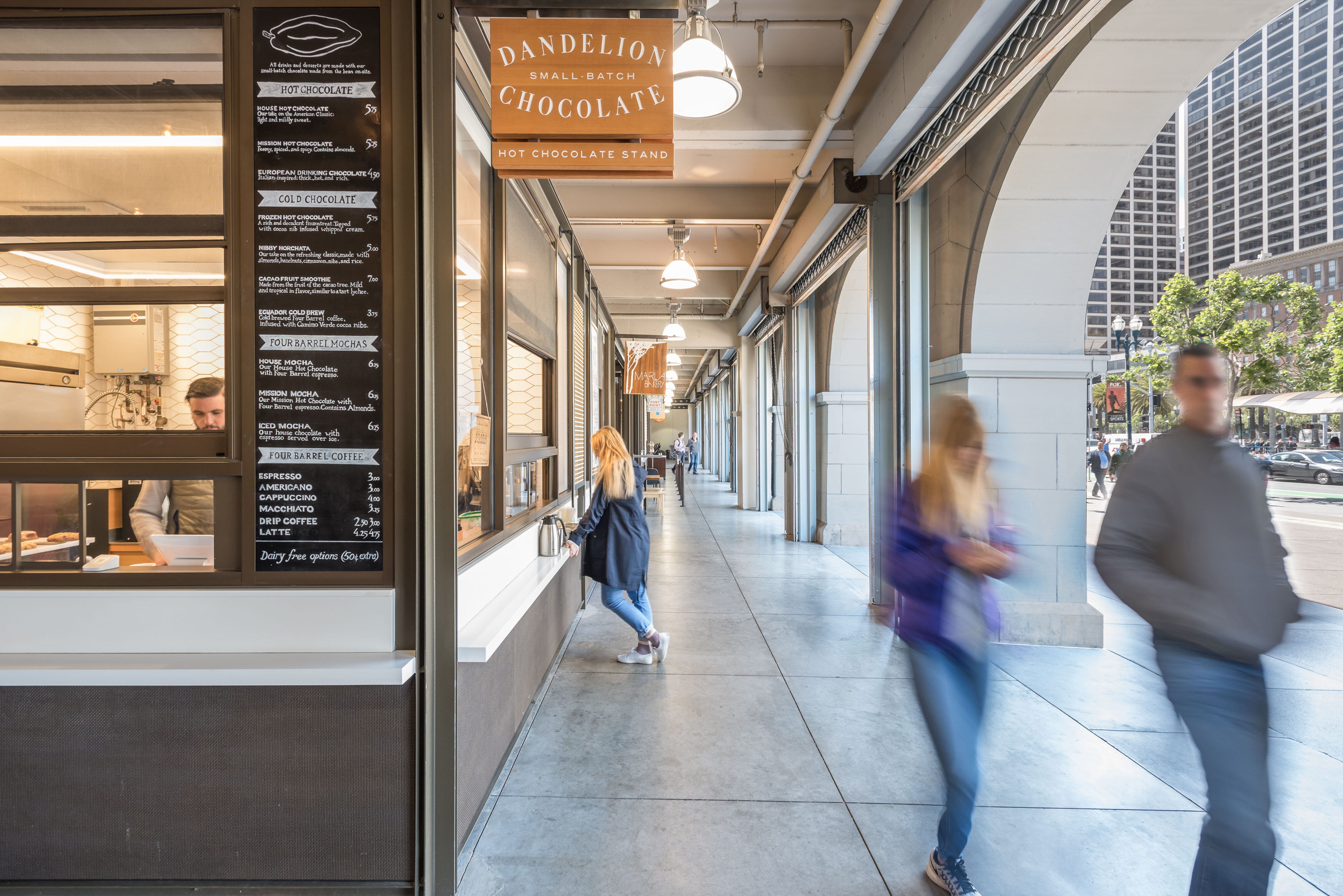Covered waterfront arcade with small retail kiosks and café windows, where people walk past a hot chocolate stand beneath arched openings overlooking a busy San Francisco street.