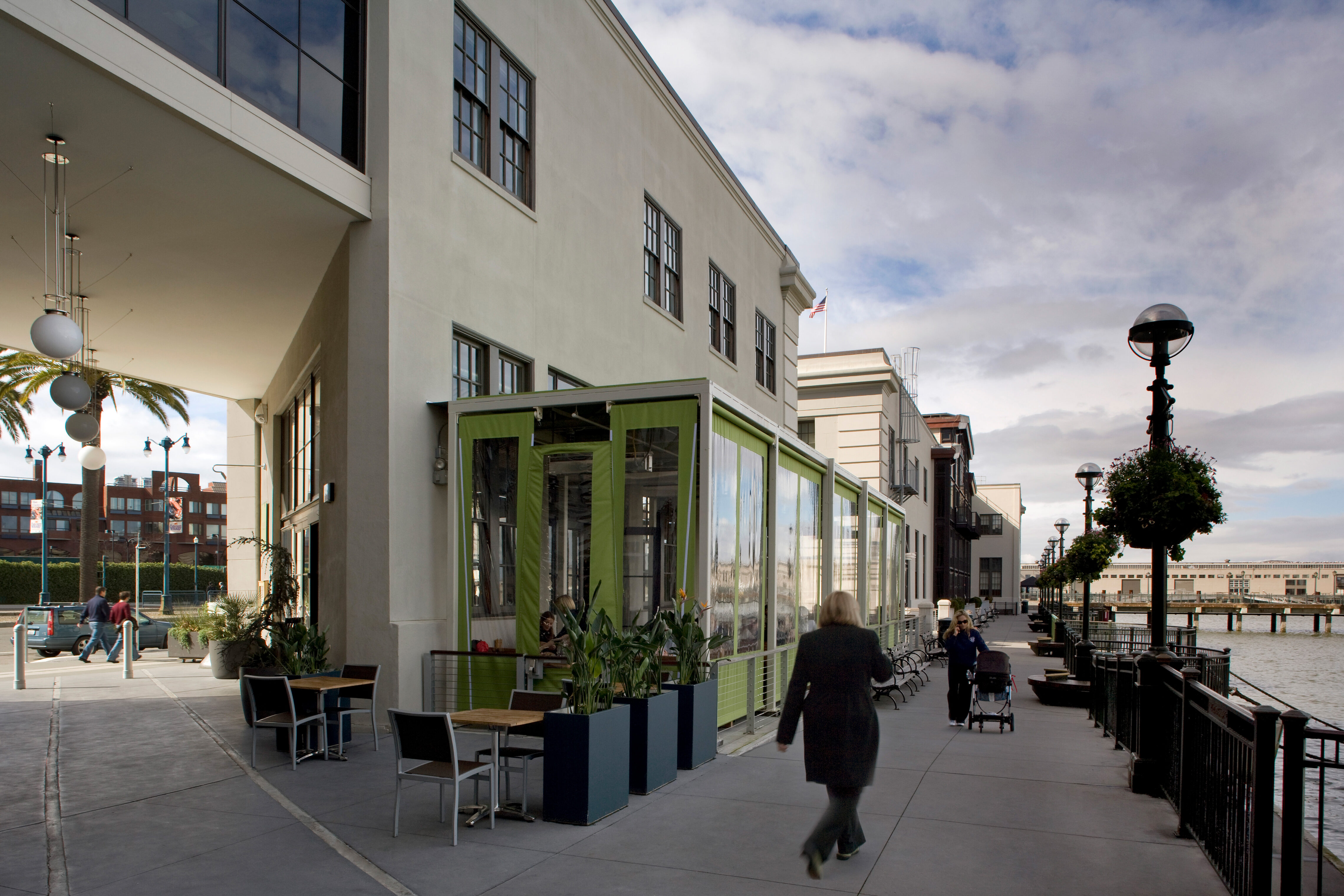 Pedestrian promenade along a renovated waterfront building with café seating and glass-enclosed dining, as people walk beside the bay under a covered arcade.