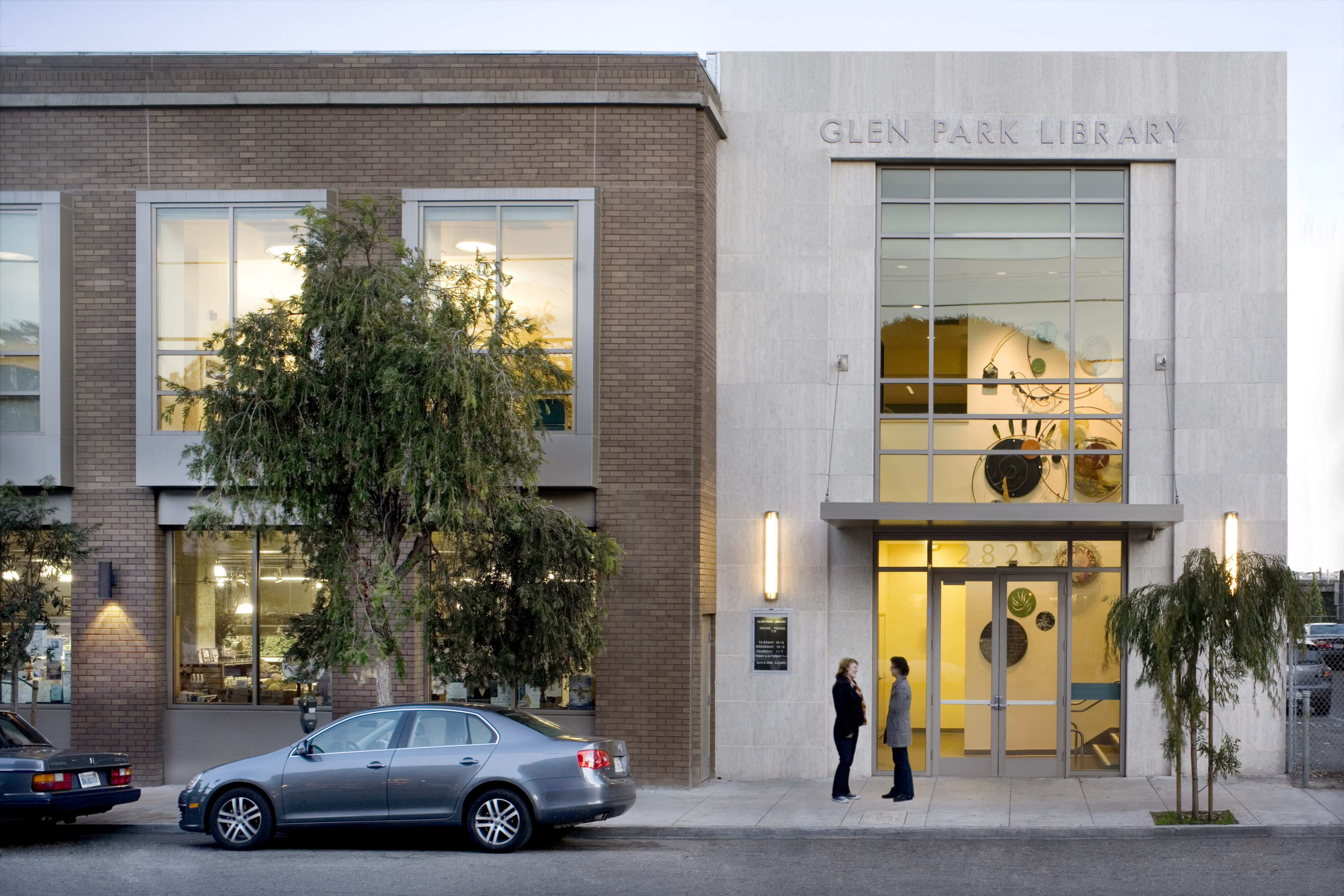 Street view of the Glen Park Library entrance at dusk, with large windows and two people standing outside.