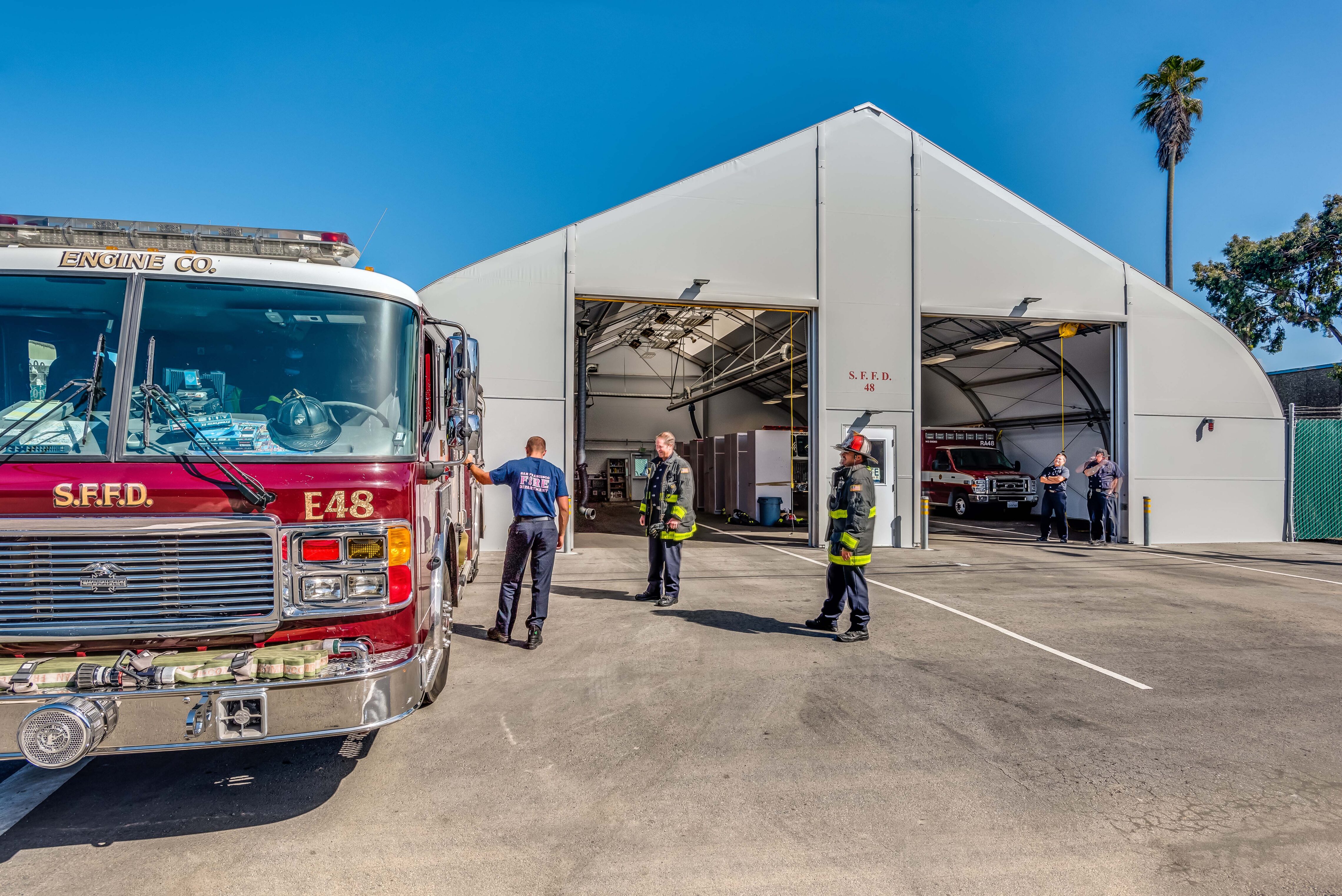 Exterior view of a fire station with open bay doors, fire engines parked outside, and firefighters standing and talking in front of the building under a clear blue sky.