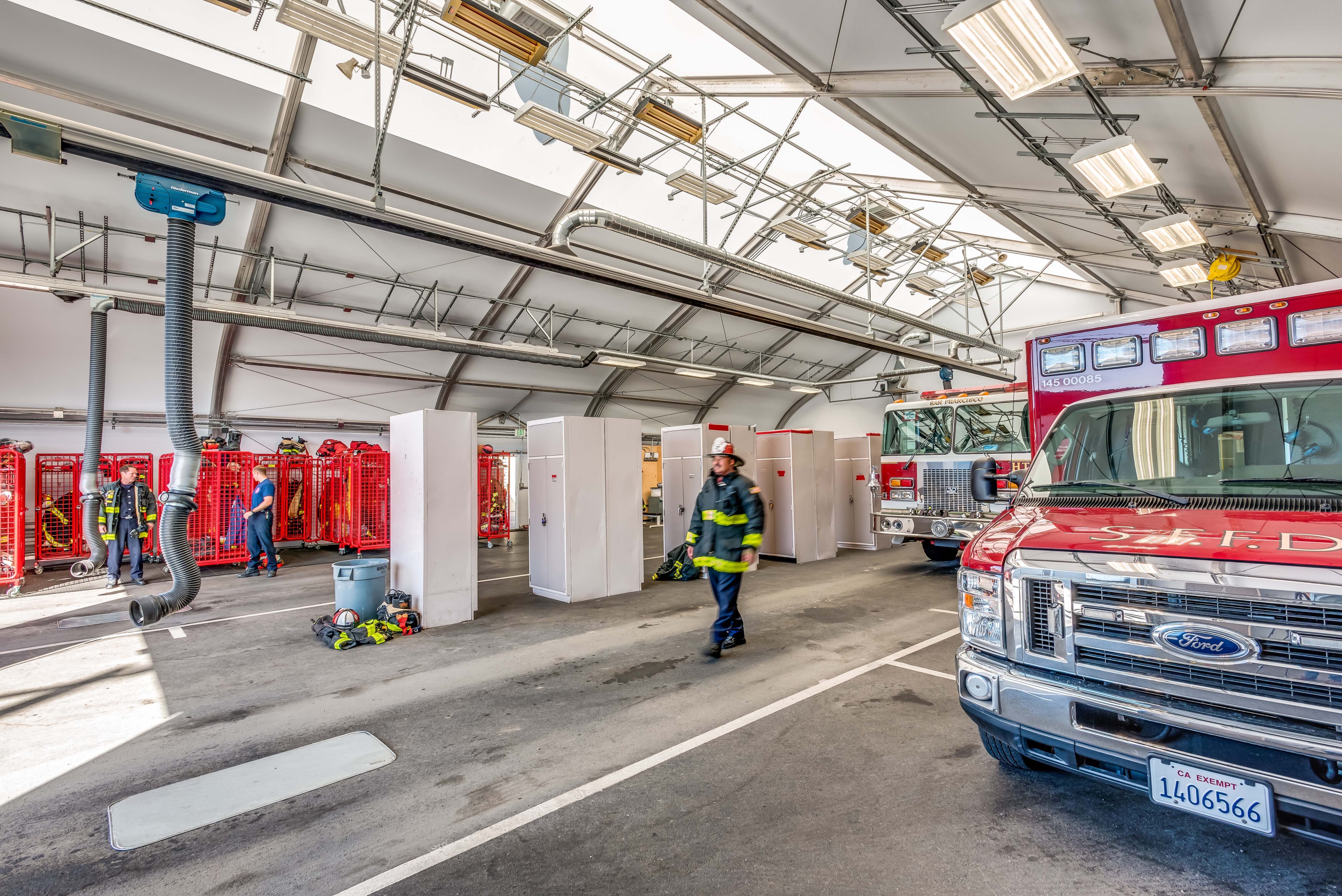 Interior of a fire station apparatus bay with fire engines parked inside, firefighters in uniform moving through the space, and overhead mechanical systems visible.