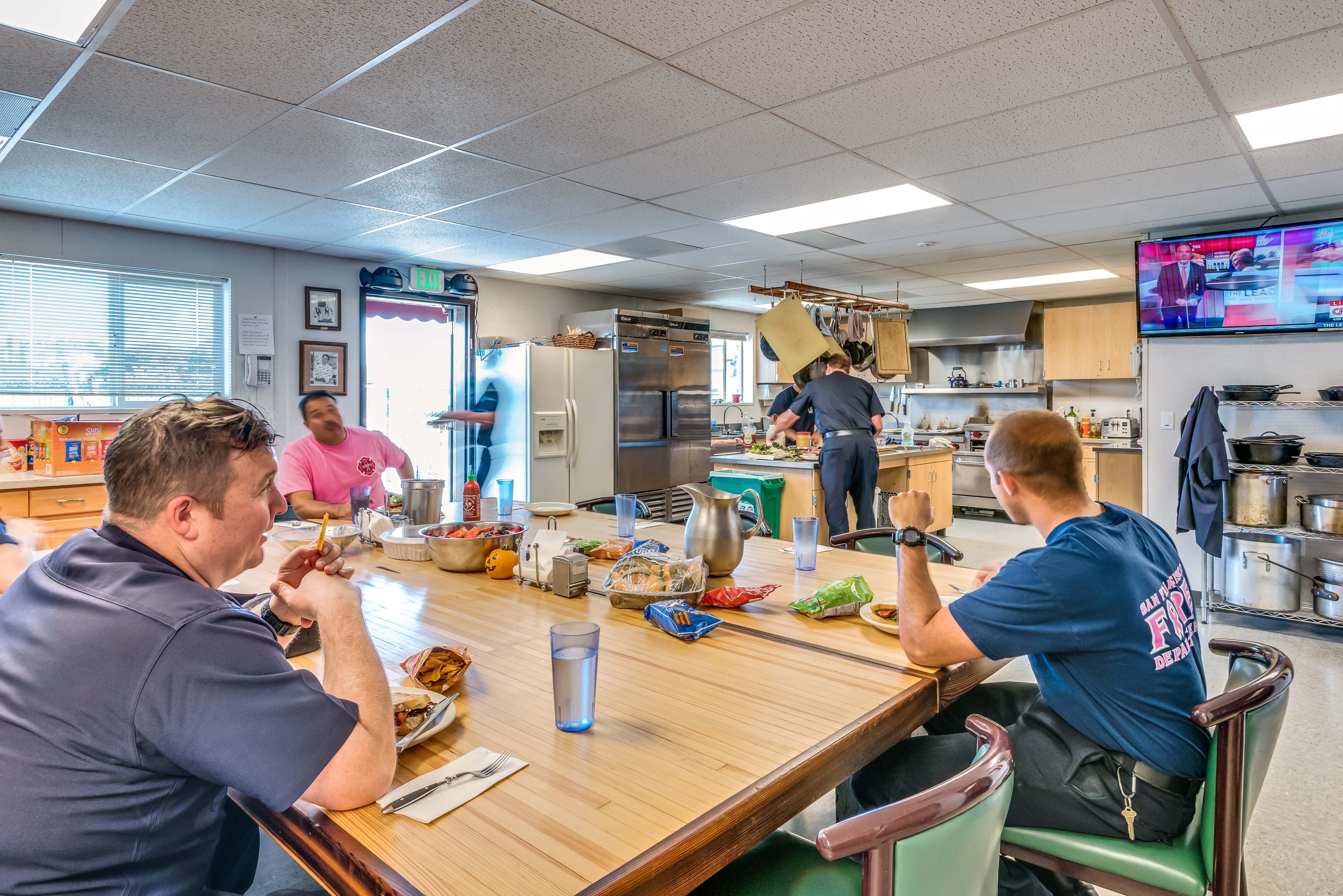 Firefighters seated around a communal kitchen table inside a fire station, eating and talking while others prepare food in the background.