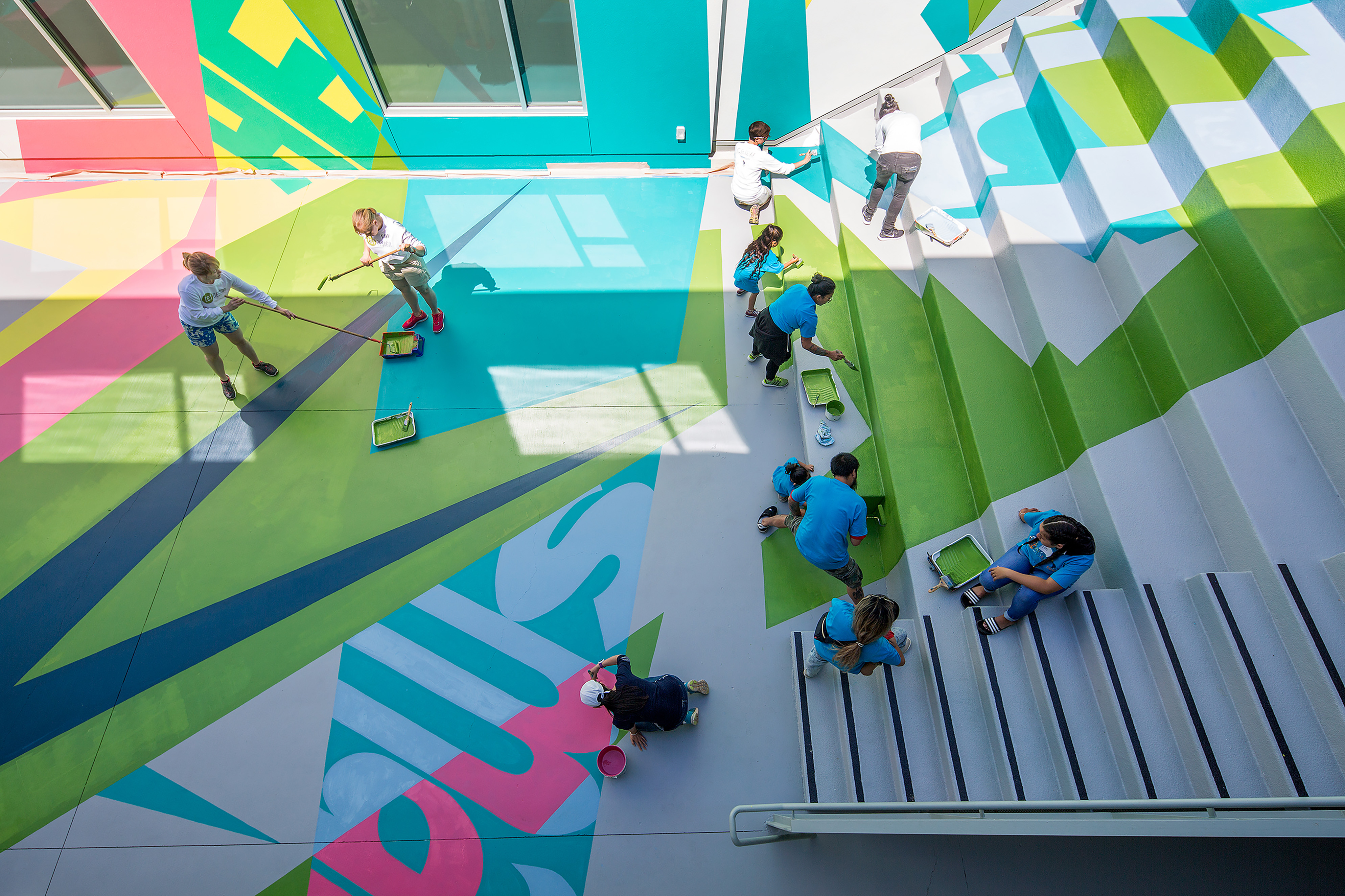TEF staff and Caliber students painting a large, colorful geometric mural across stepped seating and an adjacent plaza, viewed from above.