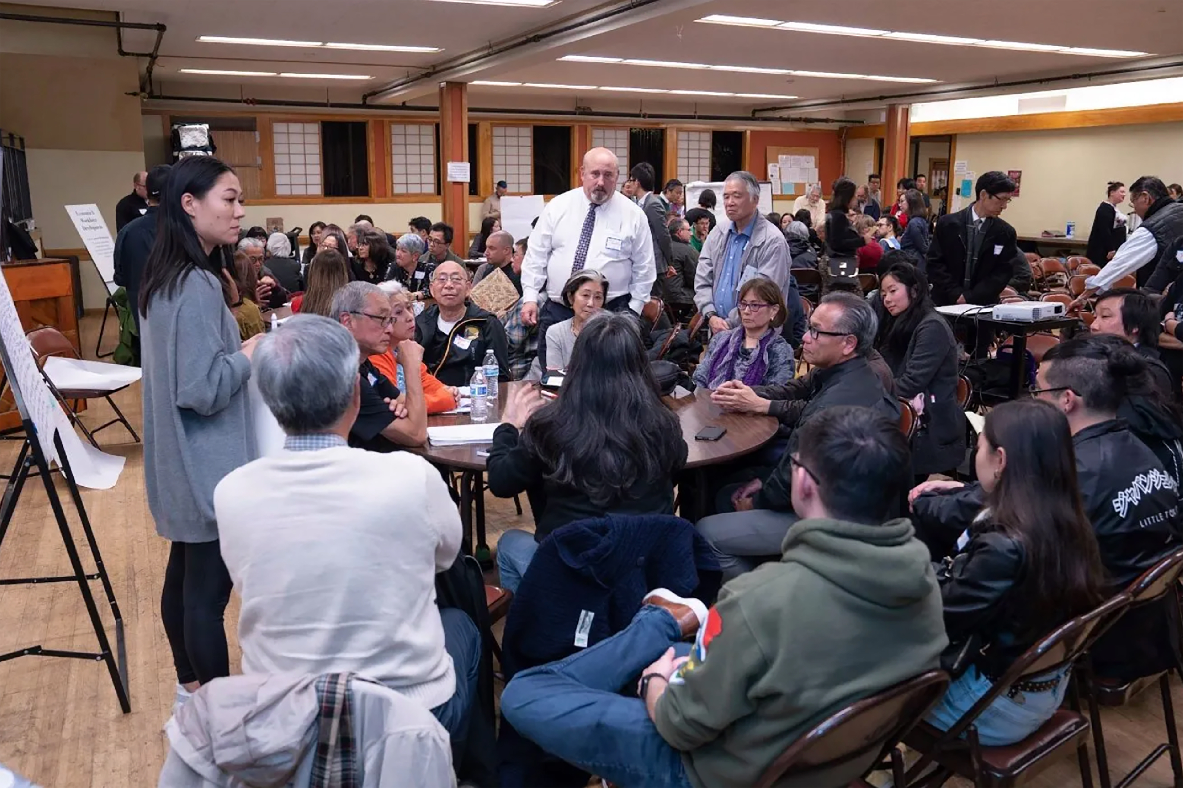 Community members gathered around tables in a large meeting hall, listening and discussing during a facilitated town hall meeting.