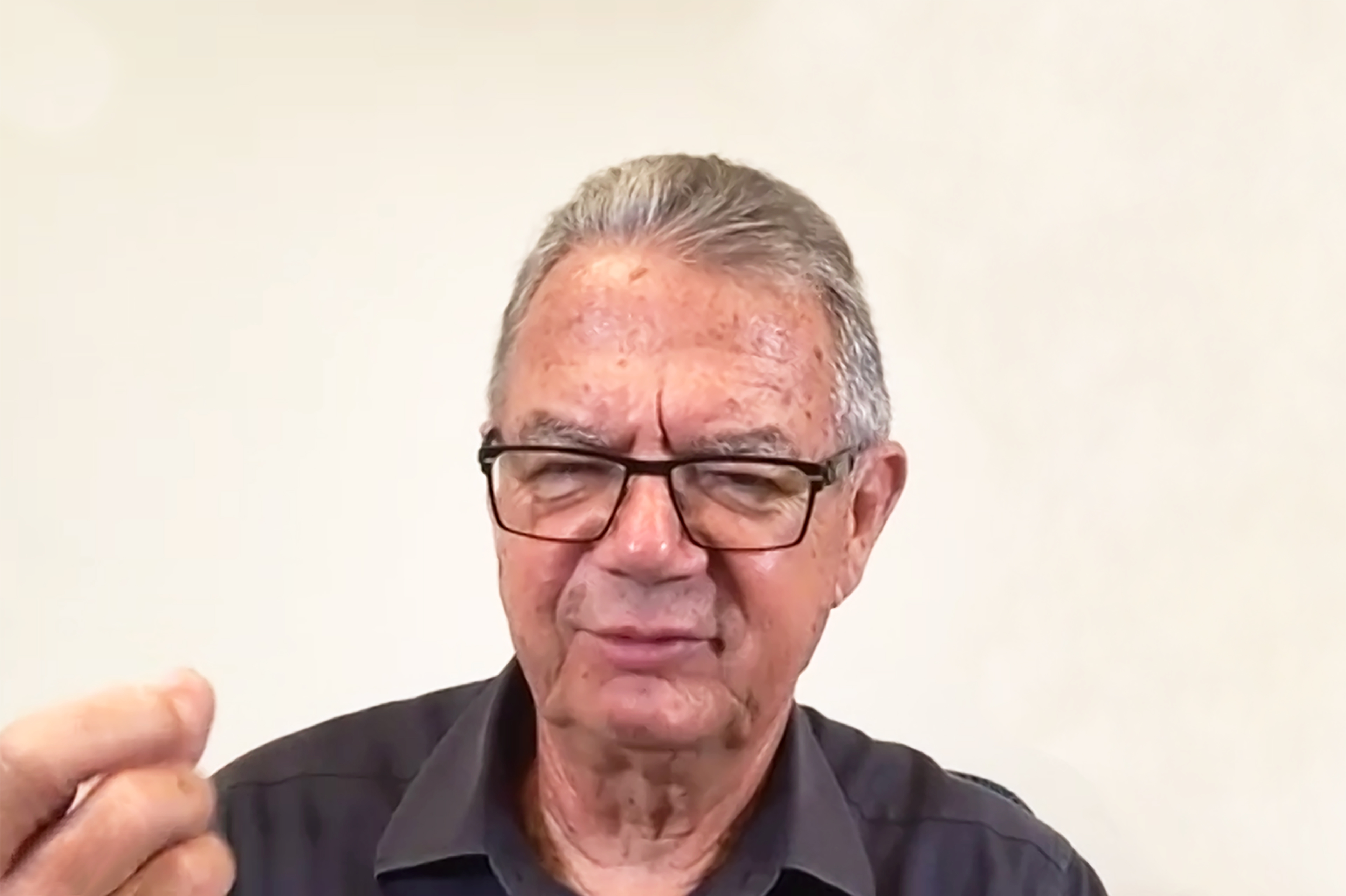 Patrick Bell with short gray hair and glasses, wearing a dark button-up shirt, speaking and gesturing with one hand against a plain light-colored background.