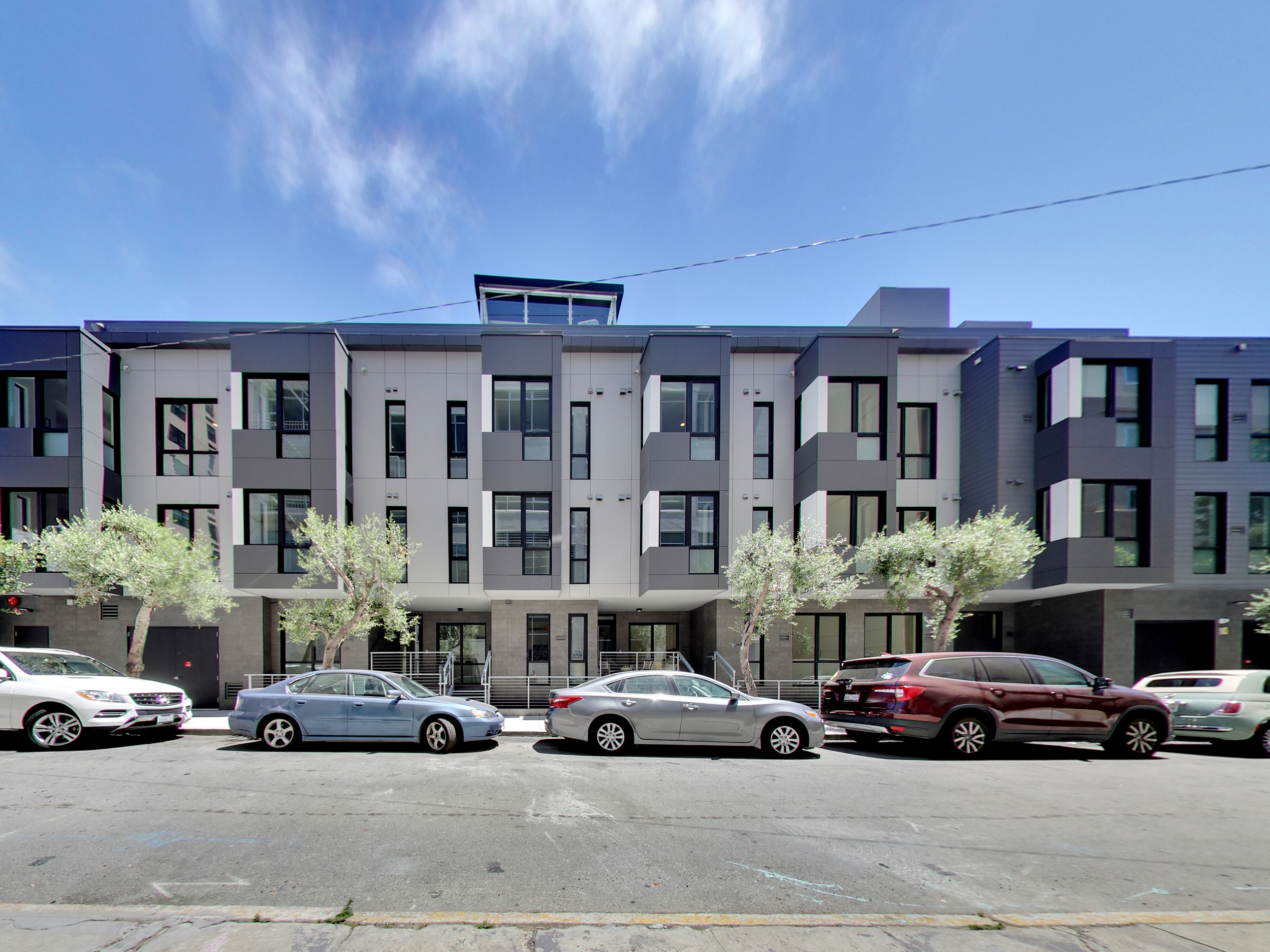 Modern four-story residential building with gray panel façades, projecting window bays, street trees, and parked cars along the curb.