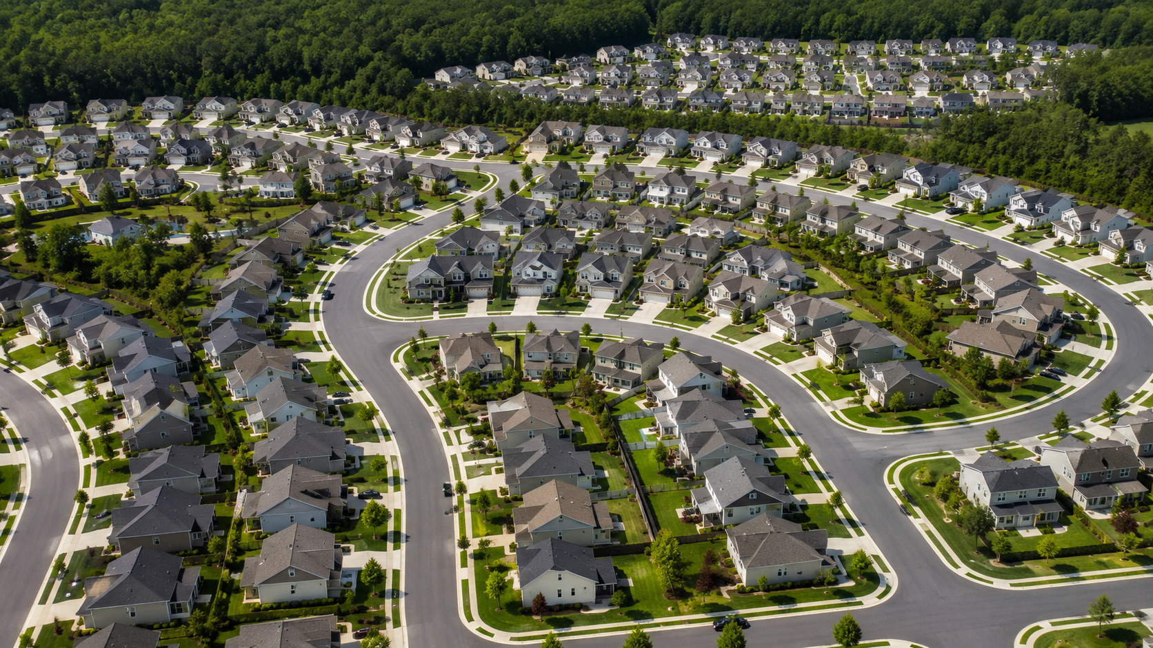Aerial view of a large suburban neighborhood with rows of similar homes arranged along curving streets and surrounded by trees.