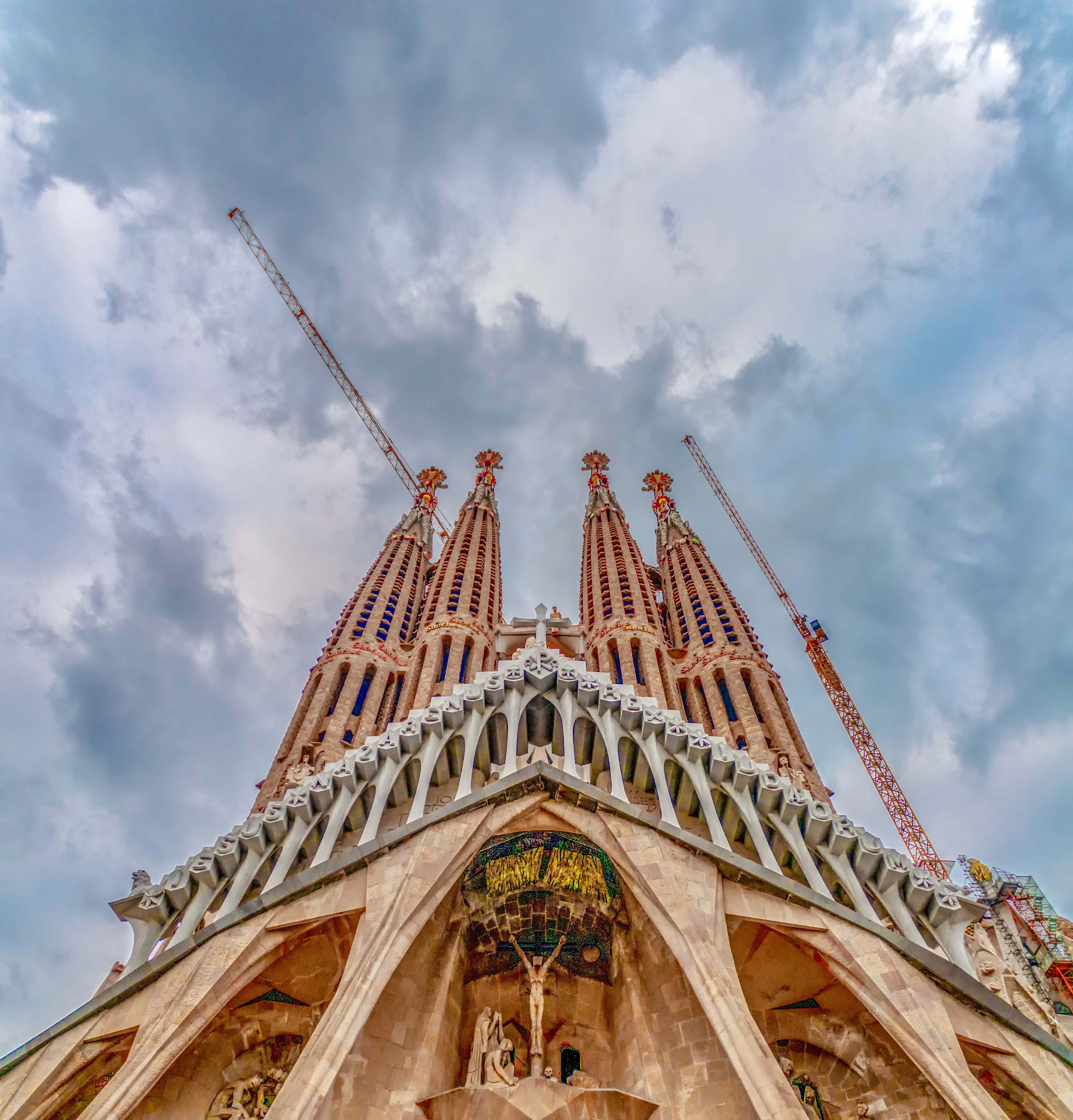 View looking up at a grand basilica with intricate stone carvings and tall spires under a cloudy sky, flanked by construction cranes.