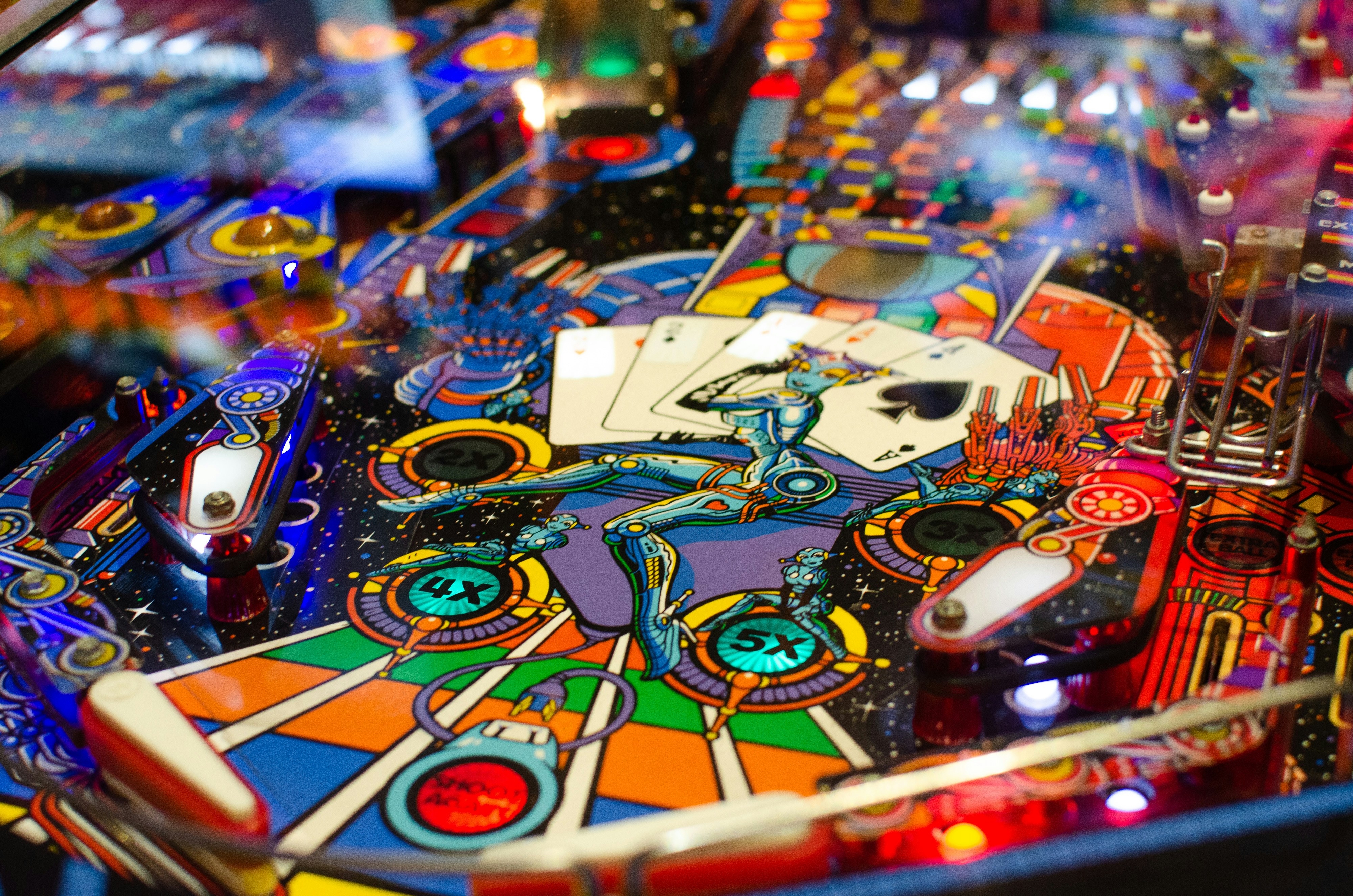 Close-up of a colorful pinball machine featuring a futuristic space-themed design with robots, planets, and playing cards illuminated by bright lights.