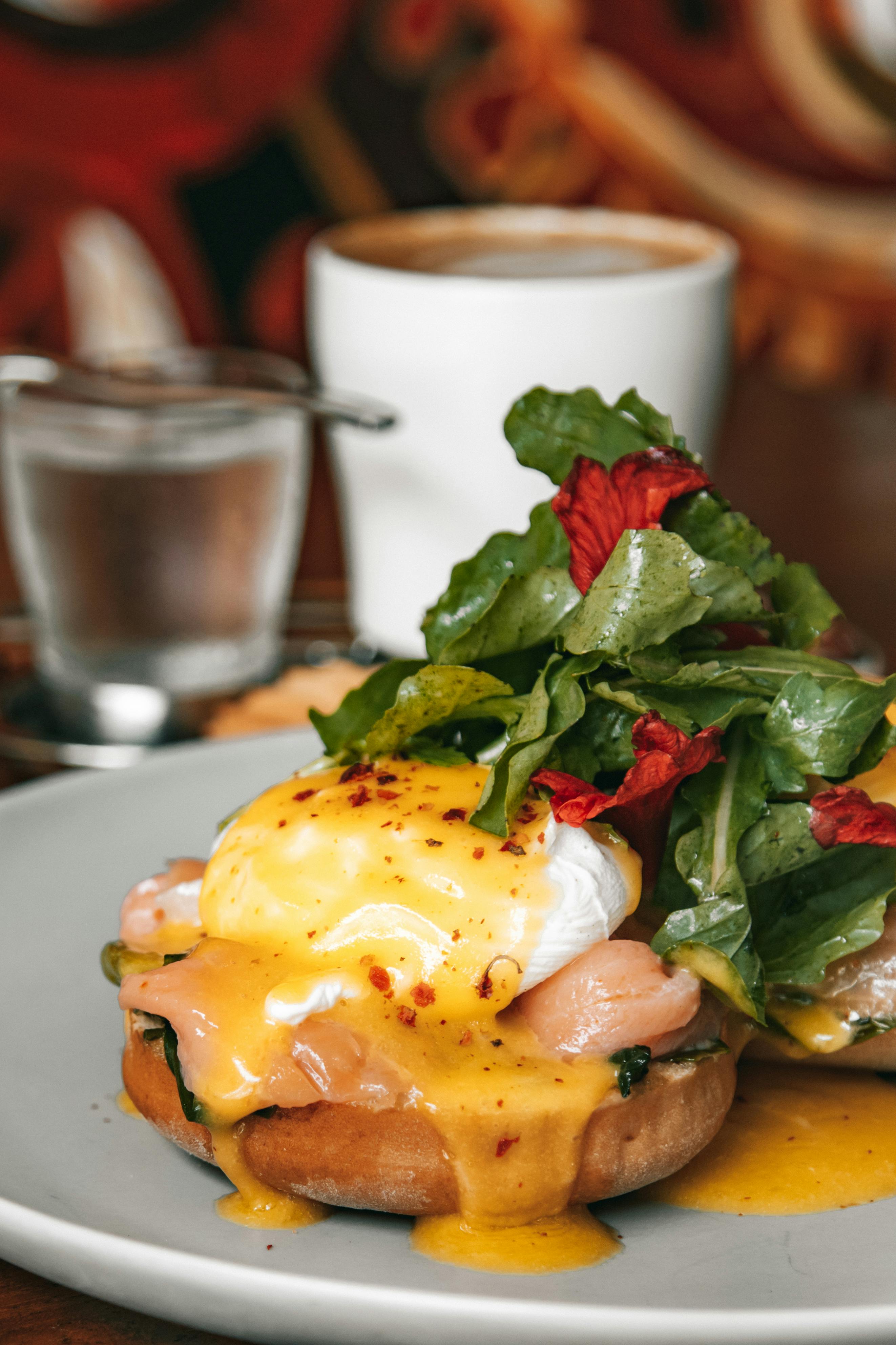 A close-up of an eggs Benedict dish topped with hollandaise sauce, smoked salmon, and fresh greens, served on a gray plate with a blurred background featuring a cup of coffee and glass of water.