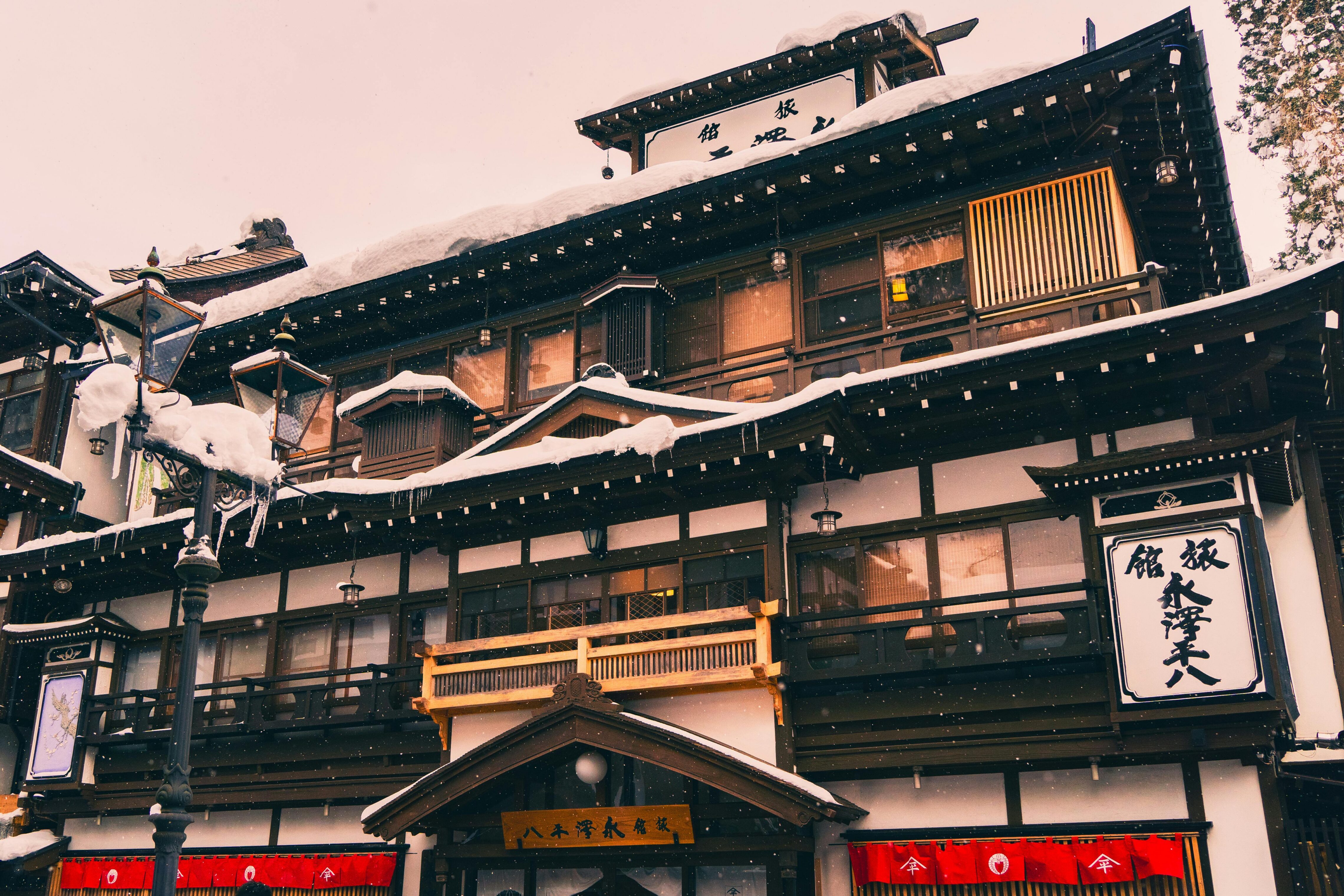 A traditional Japanese wooden hotel covered in snow, featuring dark timber framing, sliding windows, and signs with Japanese characters. Snow and icicles hang from the roof and street lamps in the foreground.