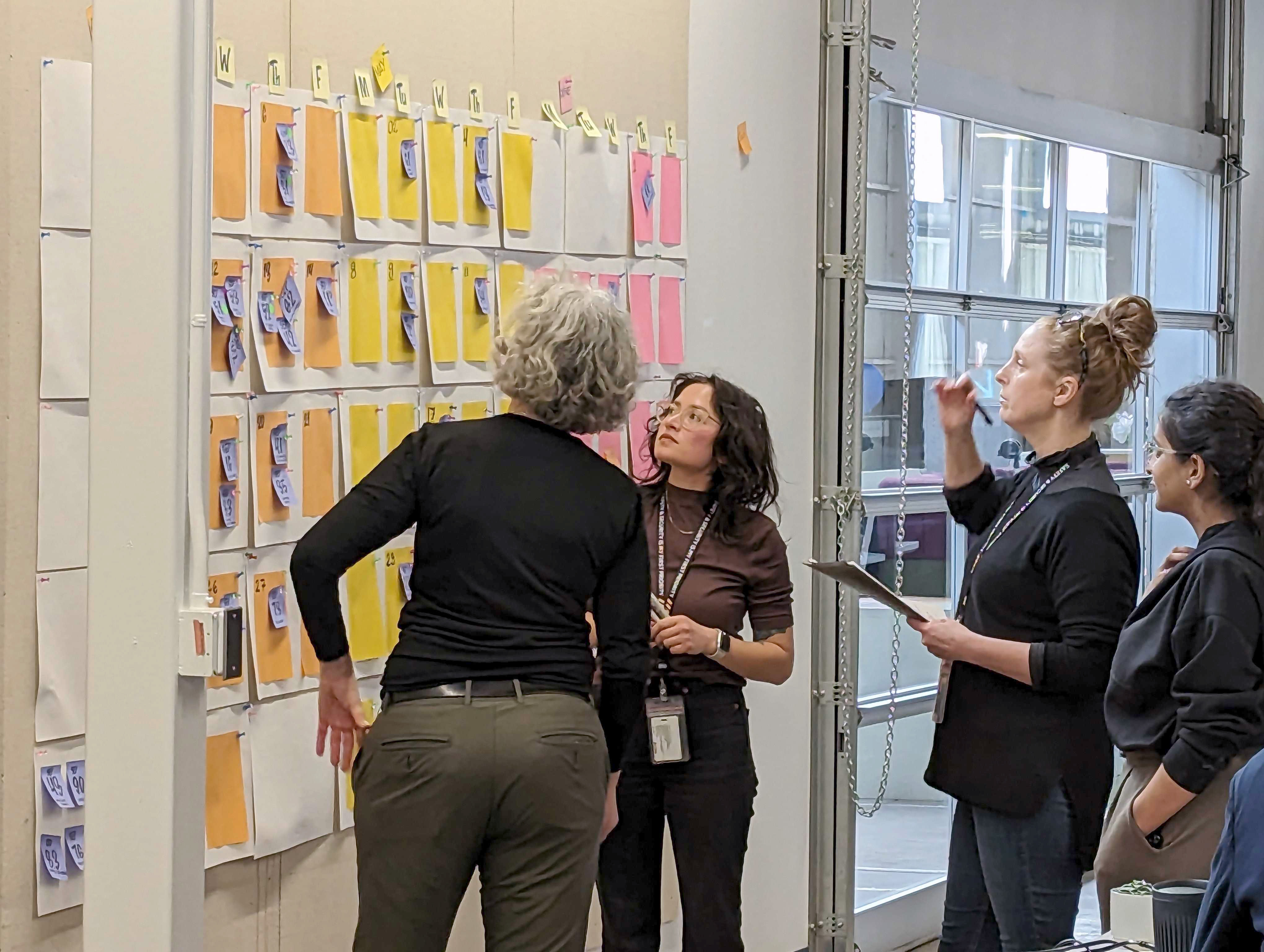 Four team members review and discuss a wall-mounted planning board covered with color-coded notes during a collaborative workshop.