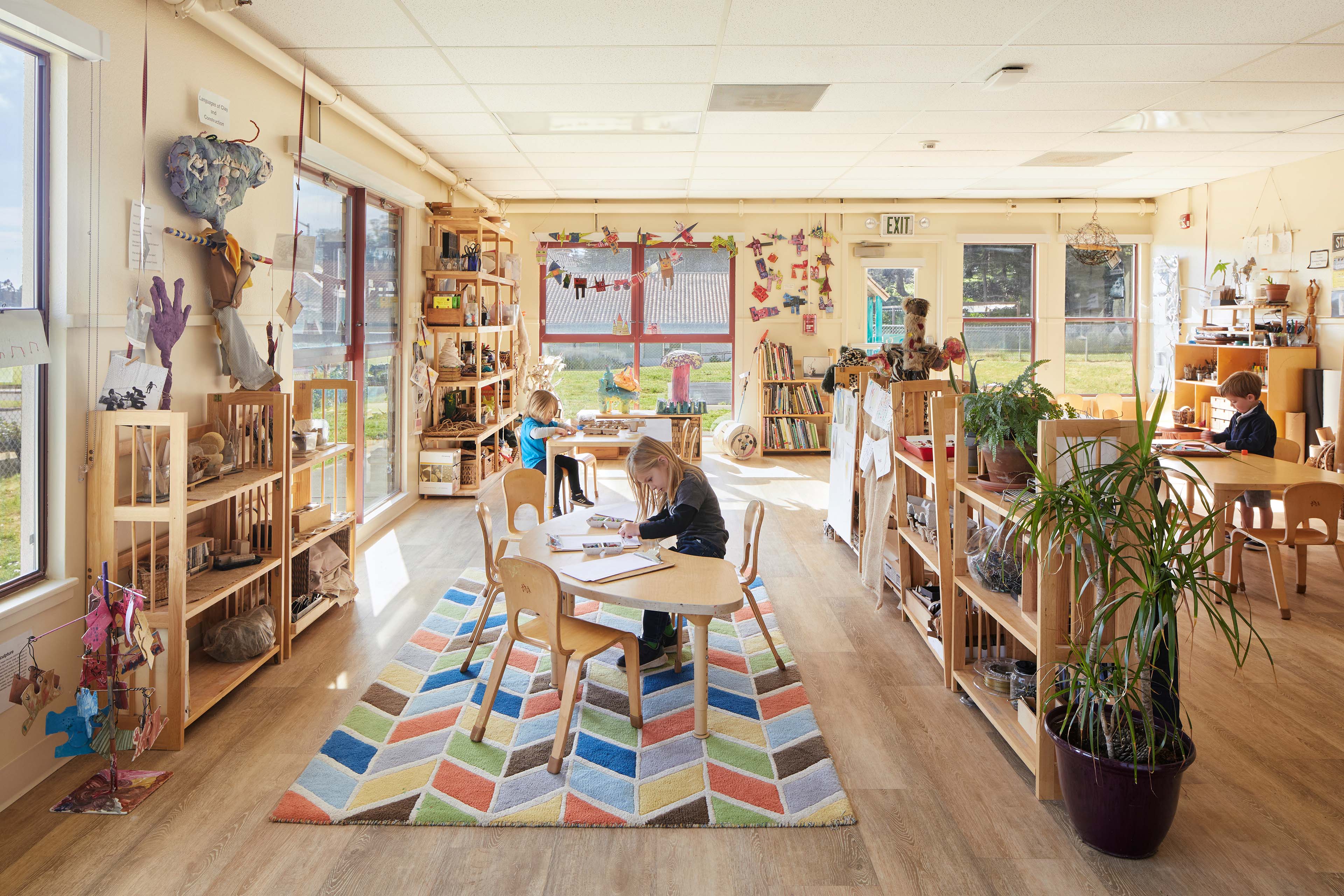 Children draw and work independently in a bright early childhood classroom with low wooden furniture, open shelving, plants, artwork, and large windows.
