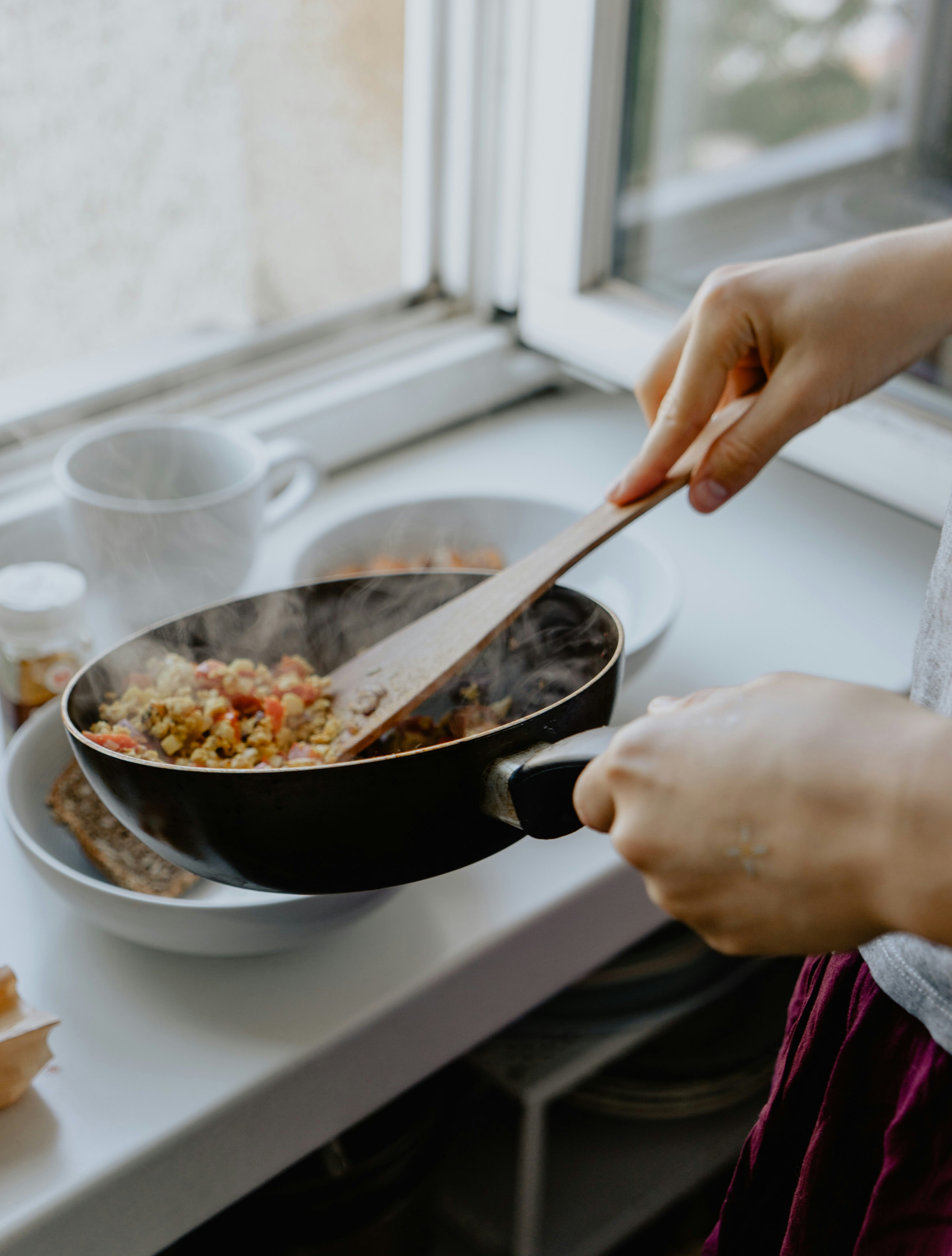 A person cooking by a bright kitchen window, stirring a steaming pan of vegetables with a wooden spoon over a countertop.