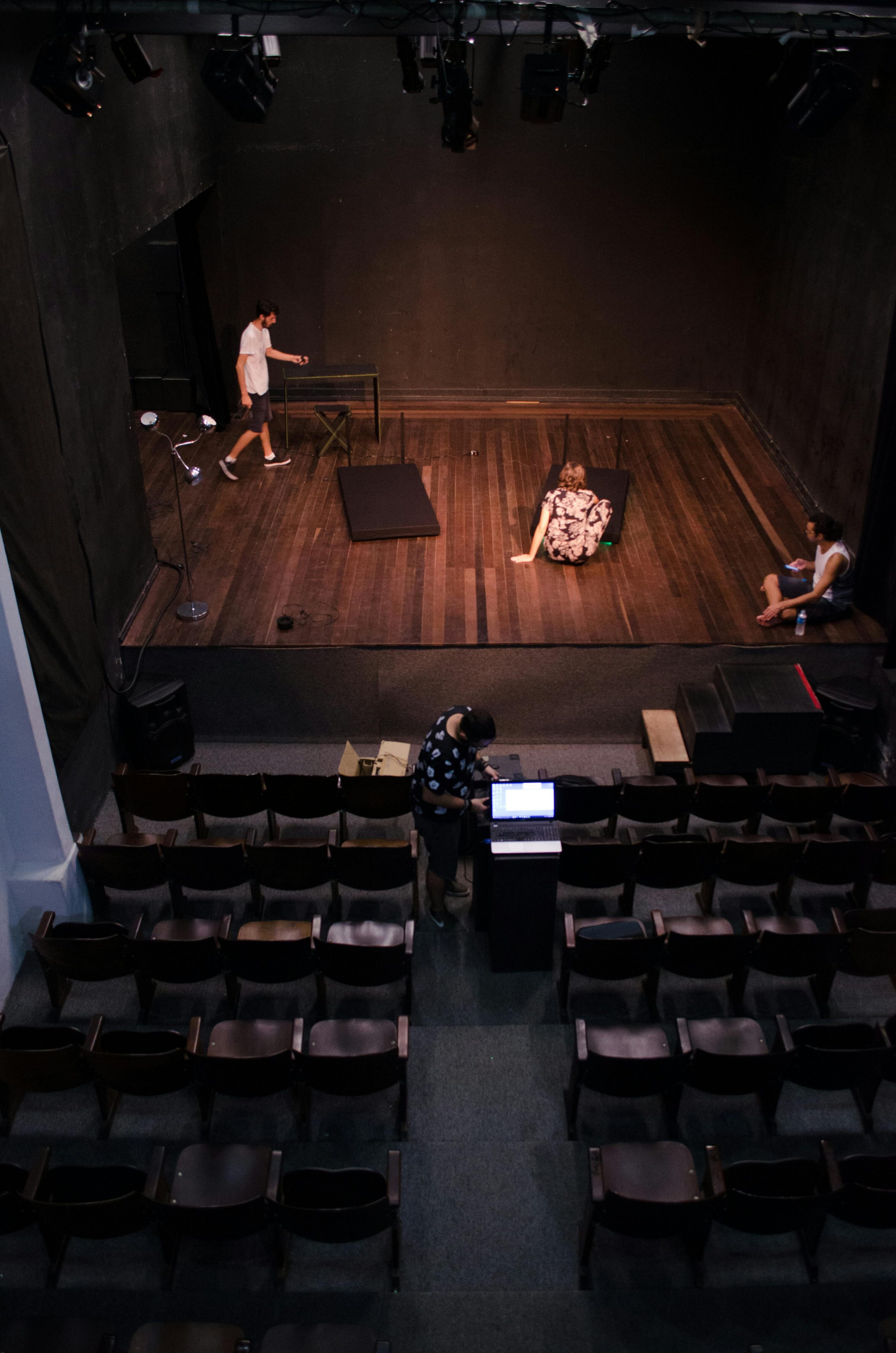 Small theater with wooden stage and rows of empty seats, where a few people are preparing equipment and rehearsing.