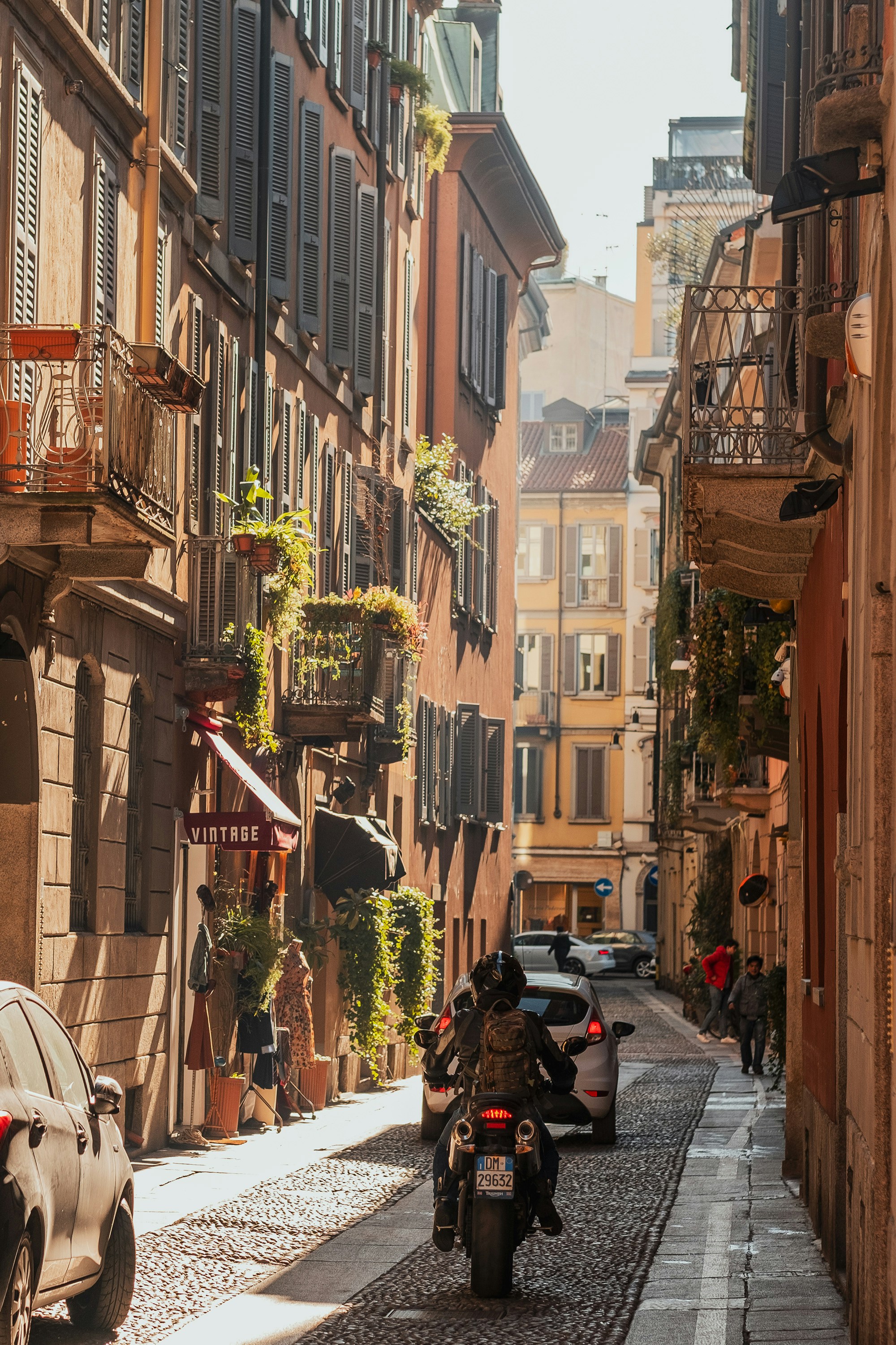 Narrow cobblestone street in Milan lined with colorful buildings, balconies with plants, and a motorcyclist riding toward the sunlight.