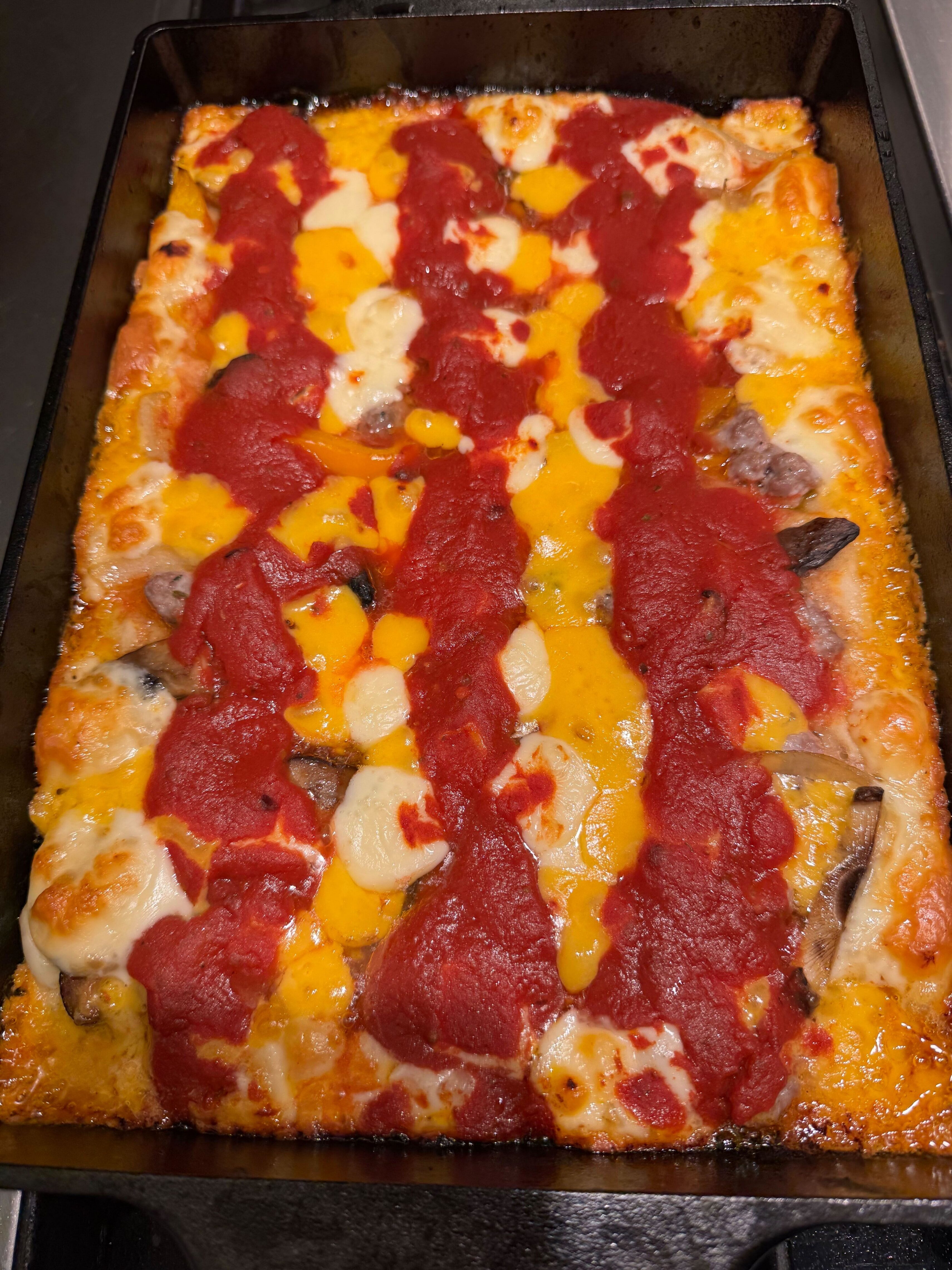 Freshly baked Detroit-style pizza in a rectangular pan, topped with melted cheese, tomato sauce stripes, and visible slices of mushrooms and peppers.