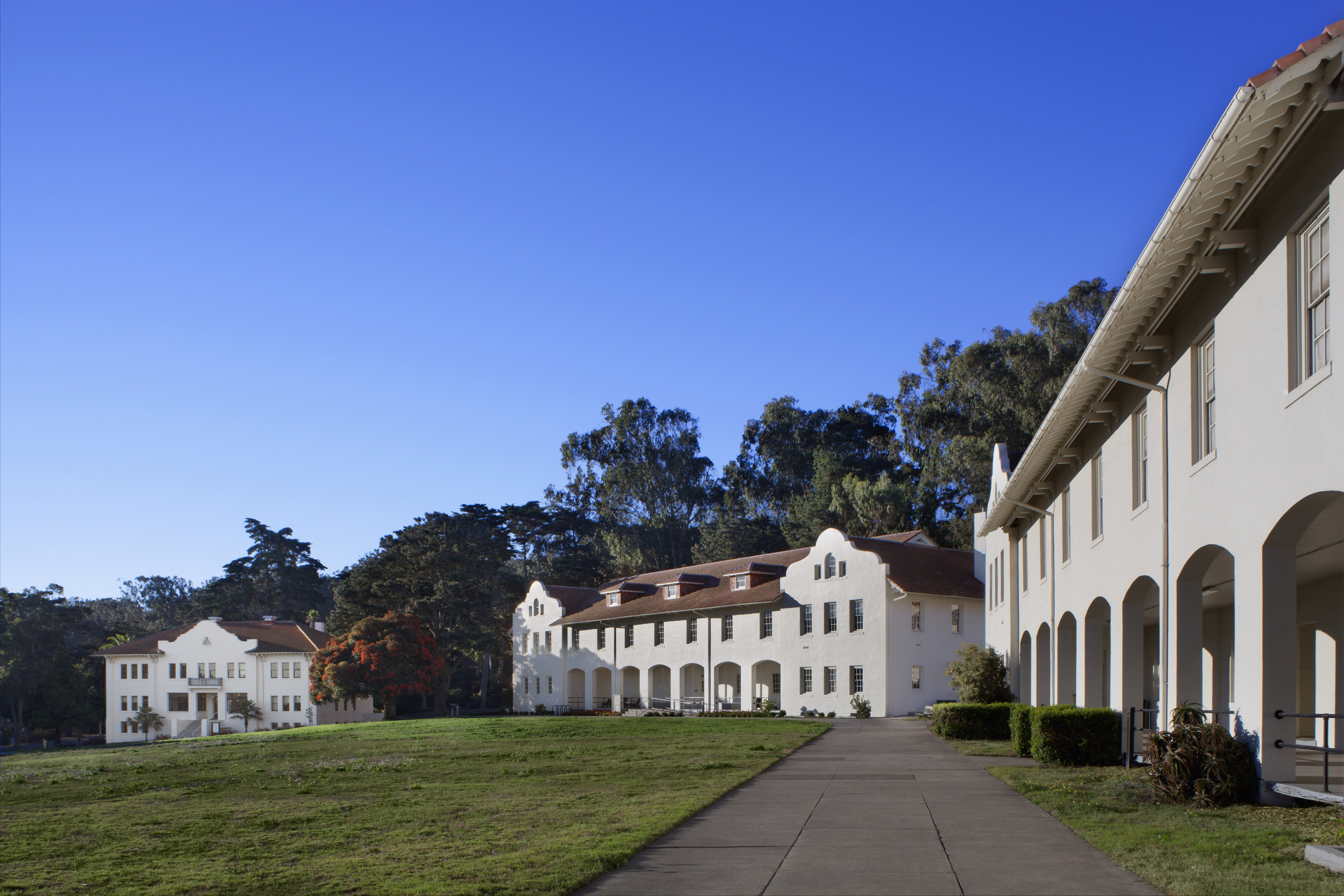 View of historic white stucco buildings with red tile roofs in the Presidio of San Francisco, set against a bright blue sky and surrounded by trees and open lawn.
