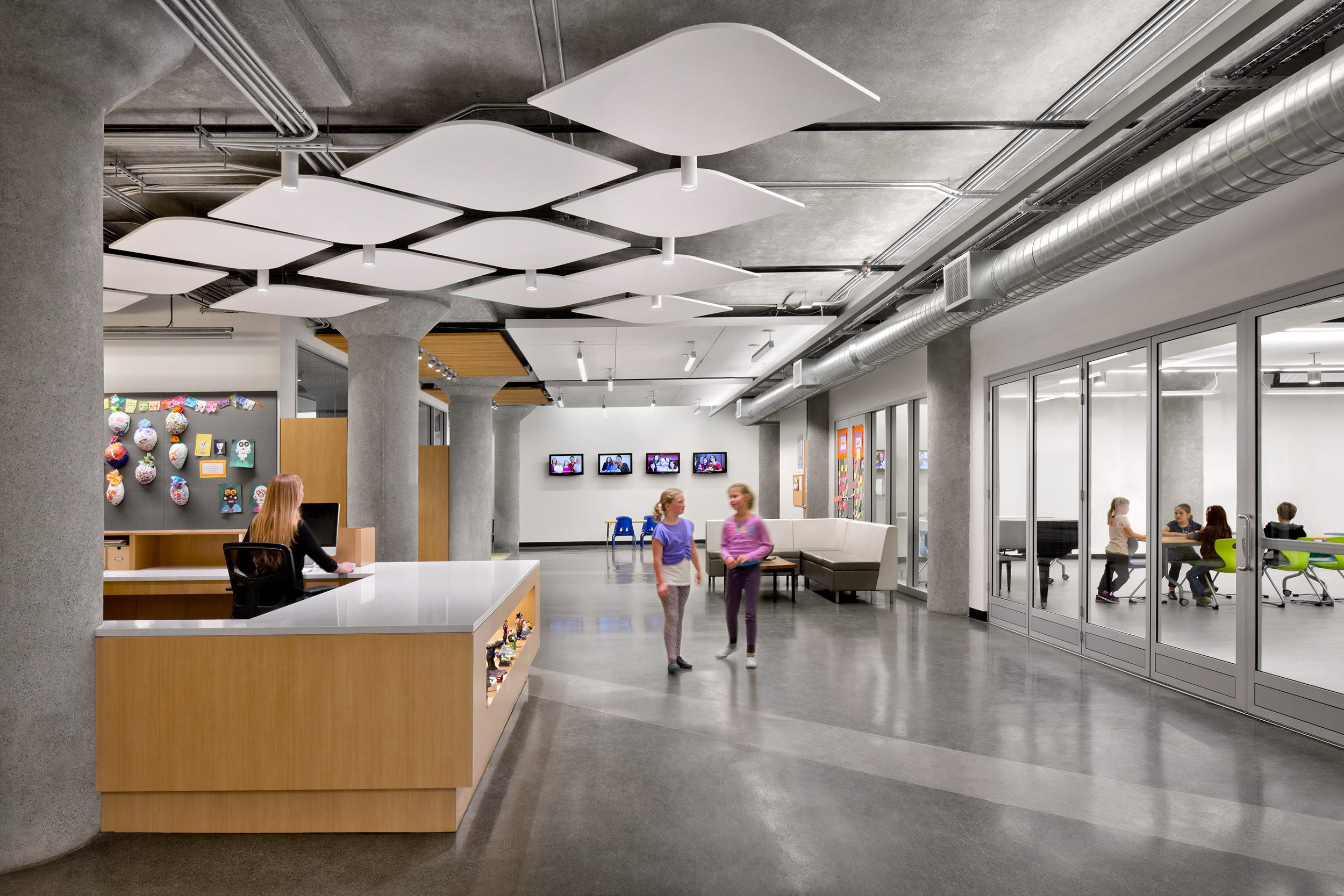Interior of the Live Oak School lobby space with concrete columns, wood finishes, and acoustic ceiling panels; two children walk past a reception desk while others work in a classroom beyond with glass walls.