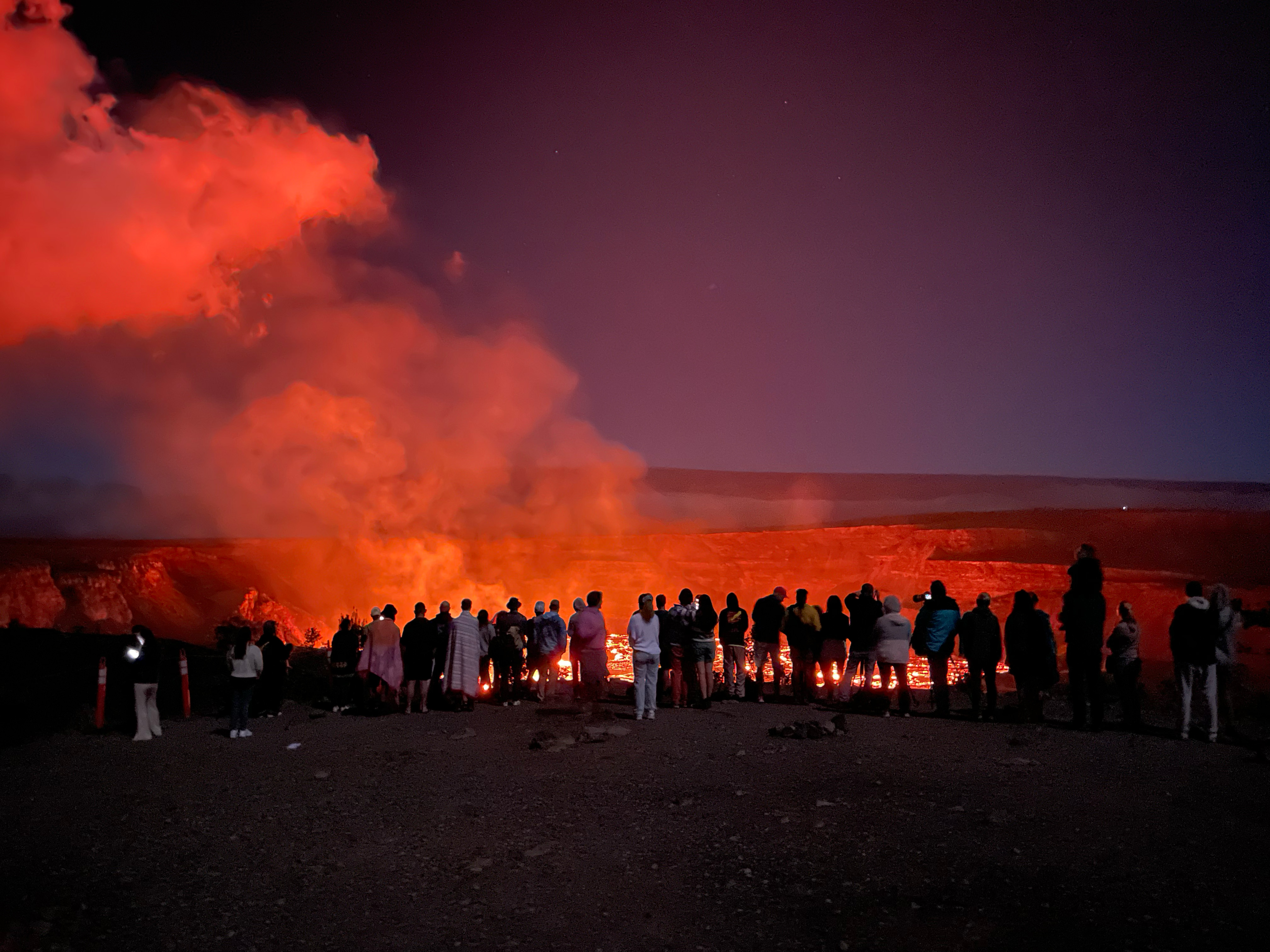 A group of people stand at night watching the glowing red lava and smoke rising from a volcanic crater. The sky above is dark blue, while the intense orange and red light from the eruption illuminates the scene, creating a dramatic contrast.