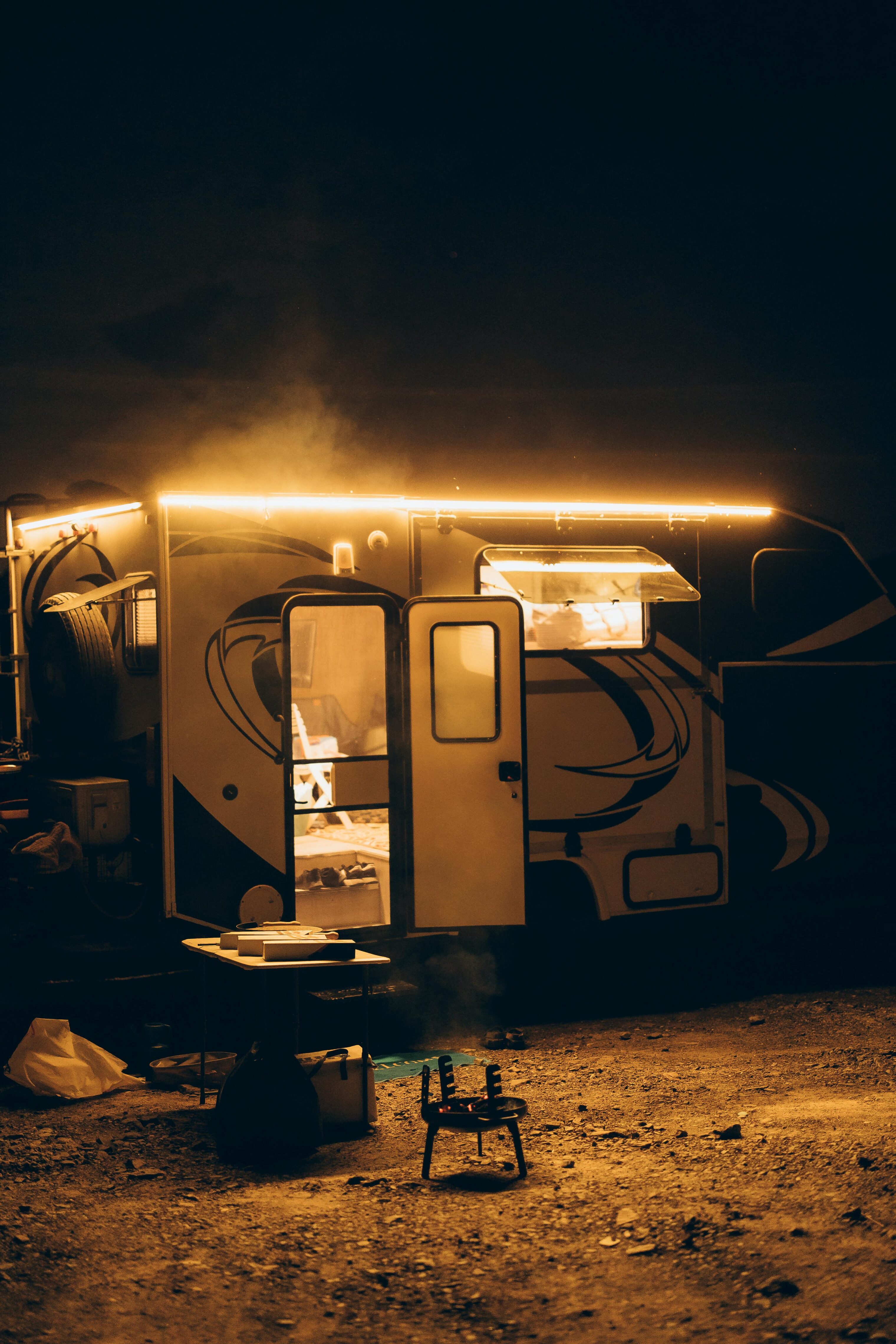 Camper trailer illuminated by warm string lights at night, with a small campfire and table set up outside on a dirt ground.