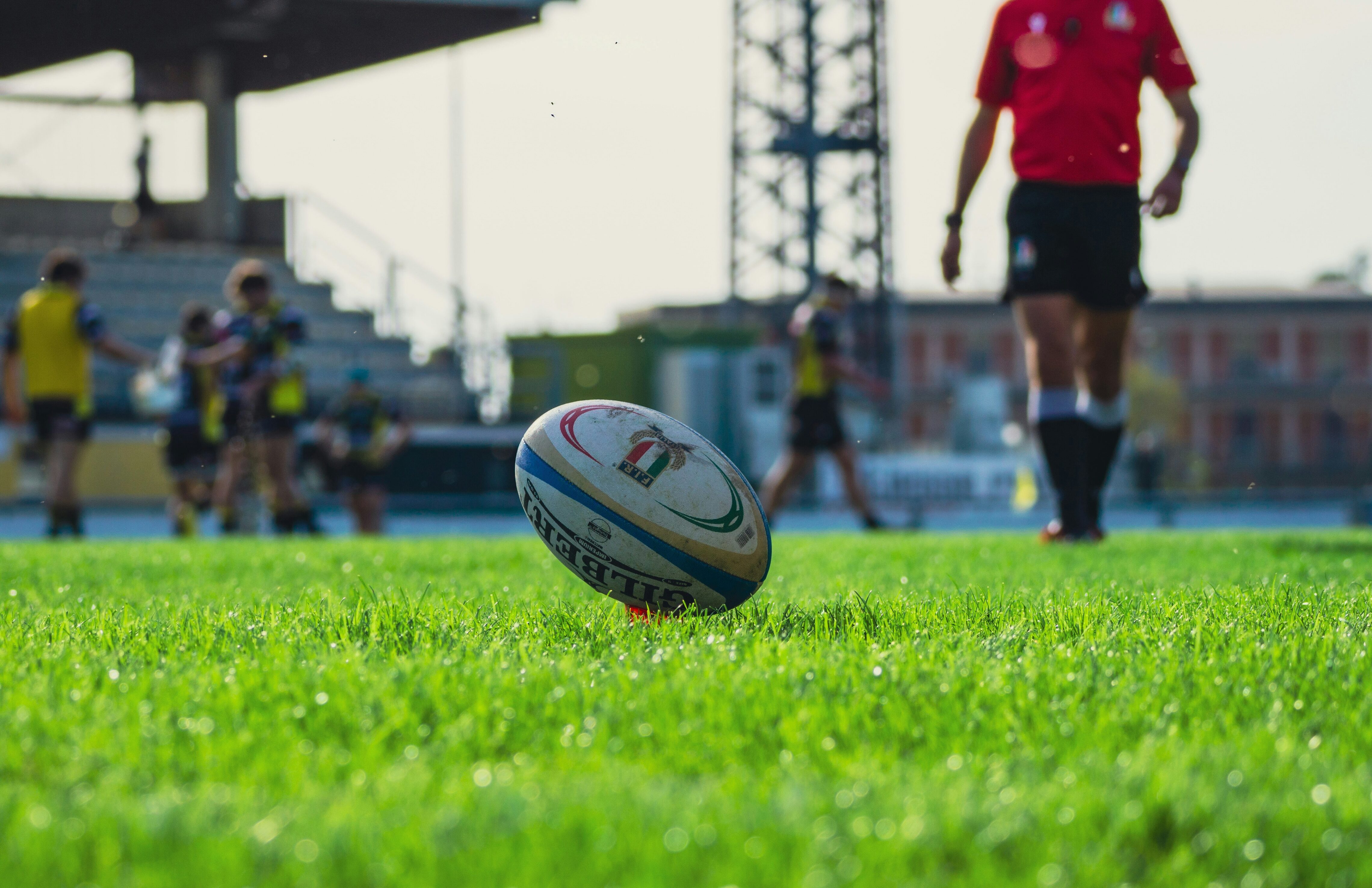 Rugby ball positioned on a grassy field with players and a referee in the background.
