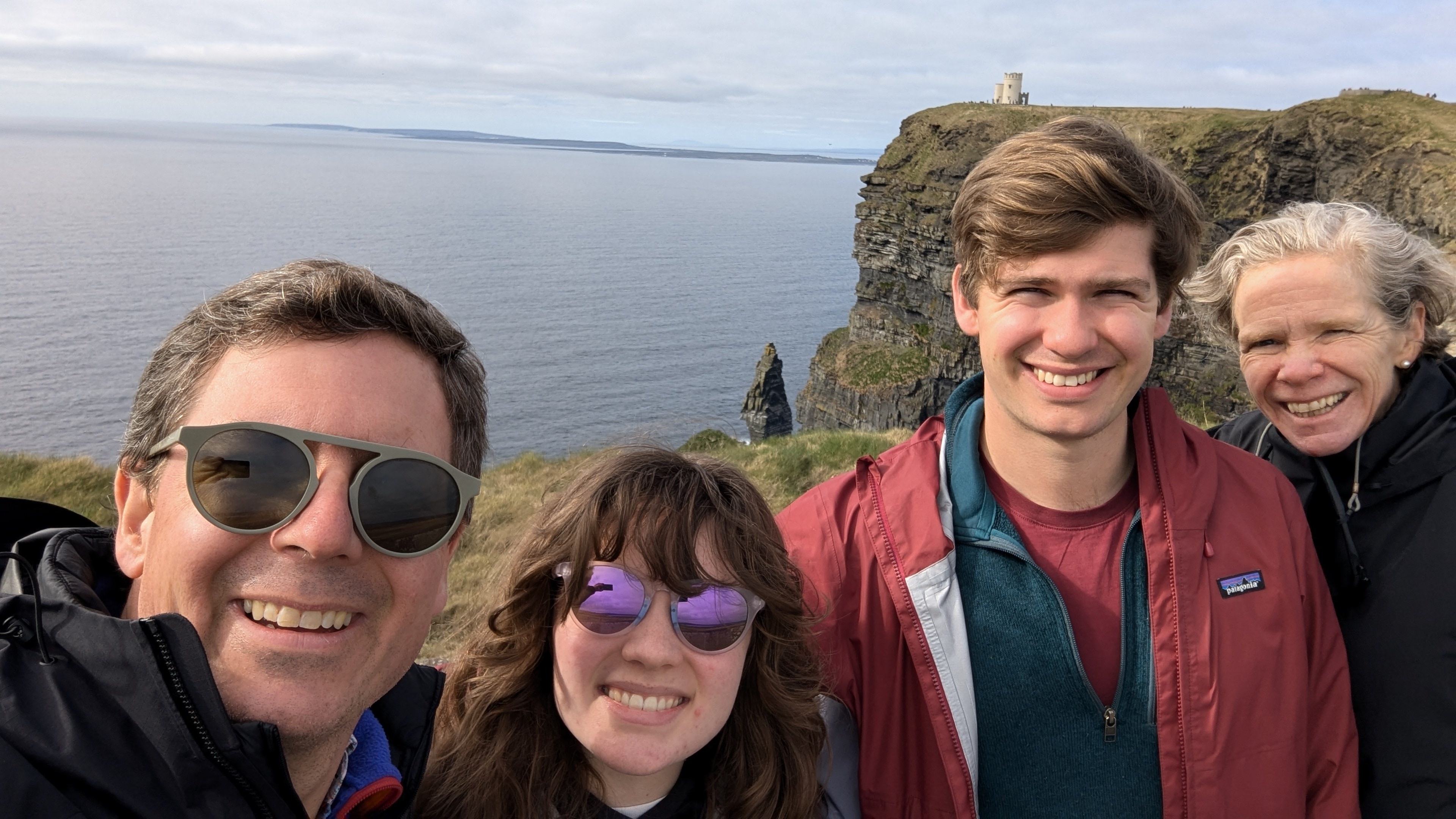 Paul Cooper with family smiling in front of ocean cliffs and a distant stone tower on a cloudy day.