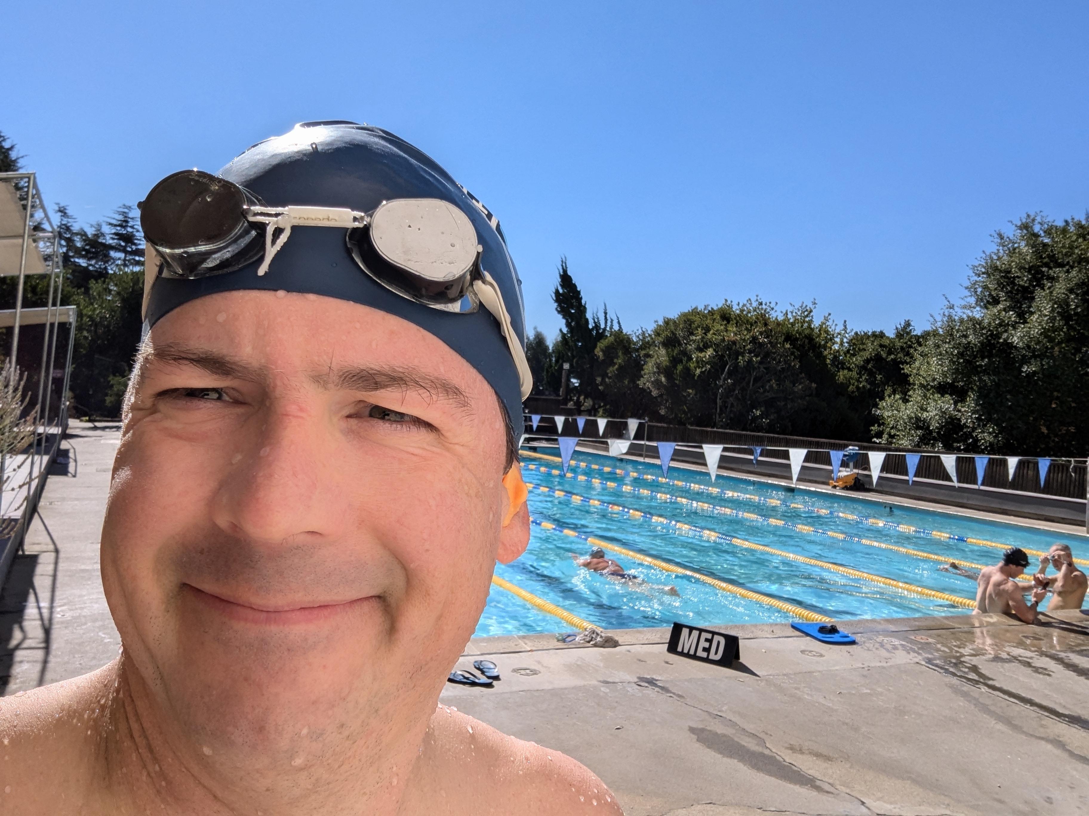 Paul Cooper smiling in swim goggles and cap beside an outdoor lap pool under clear blue skies, with other swimmers in the background.