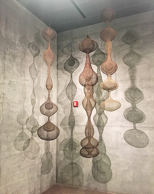 Several suspended woven-wire sculptures by Ruth Asawa cast intricate, elongated shadows across concrete gallery walls.