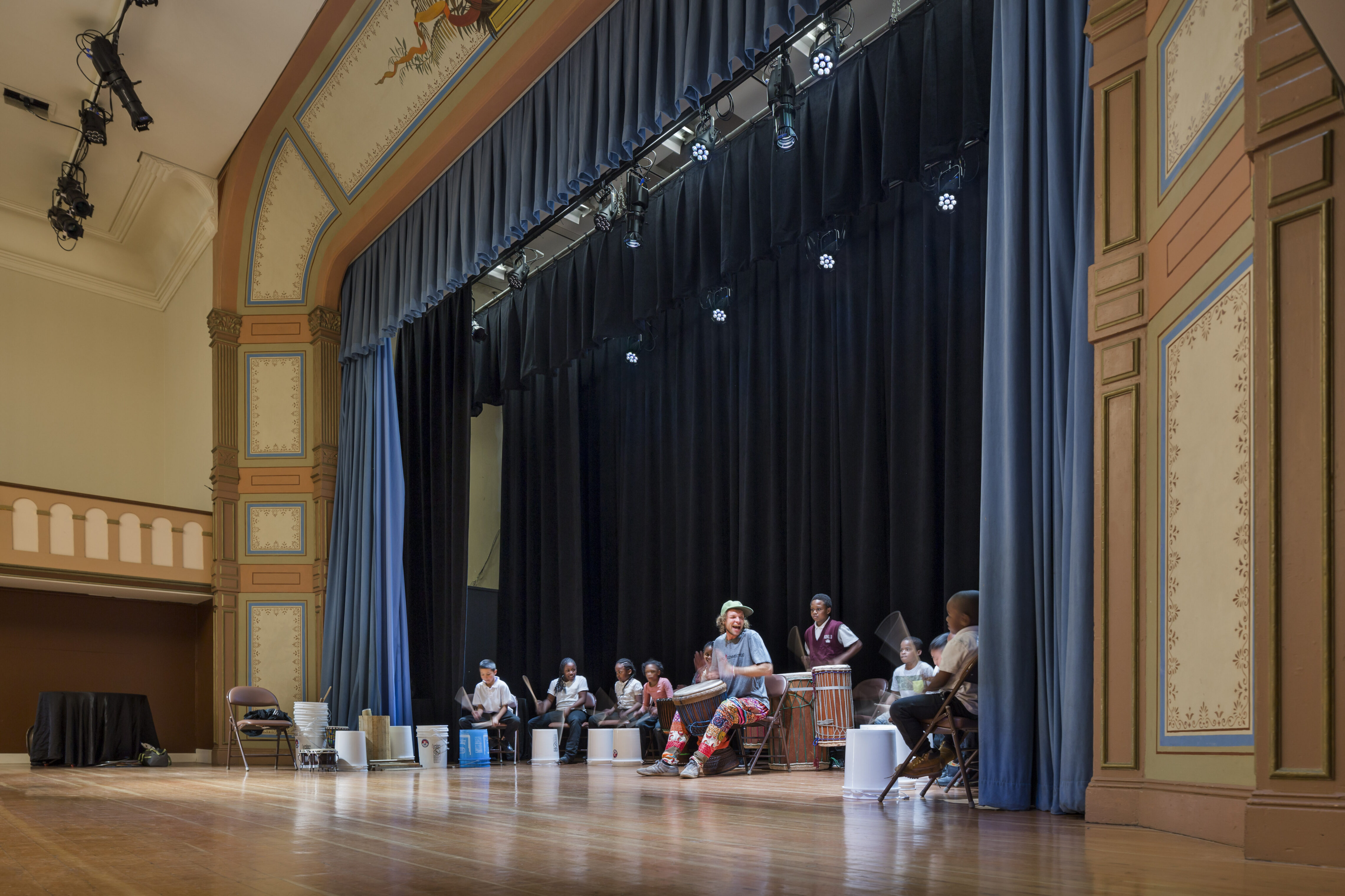 A group of children and a teacher perform a lively drumming activity on stage in a historic theater. The ornate proscenium arch features painted details and blue curtains, while stage lights illuminate the performers against a dark backdrop.
