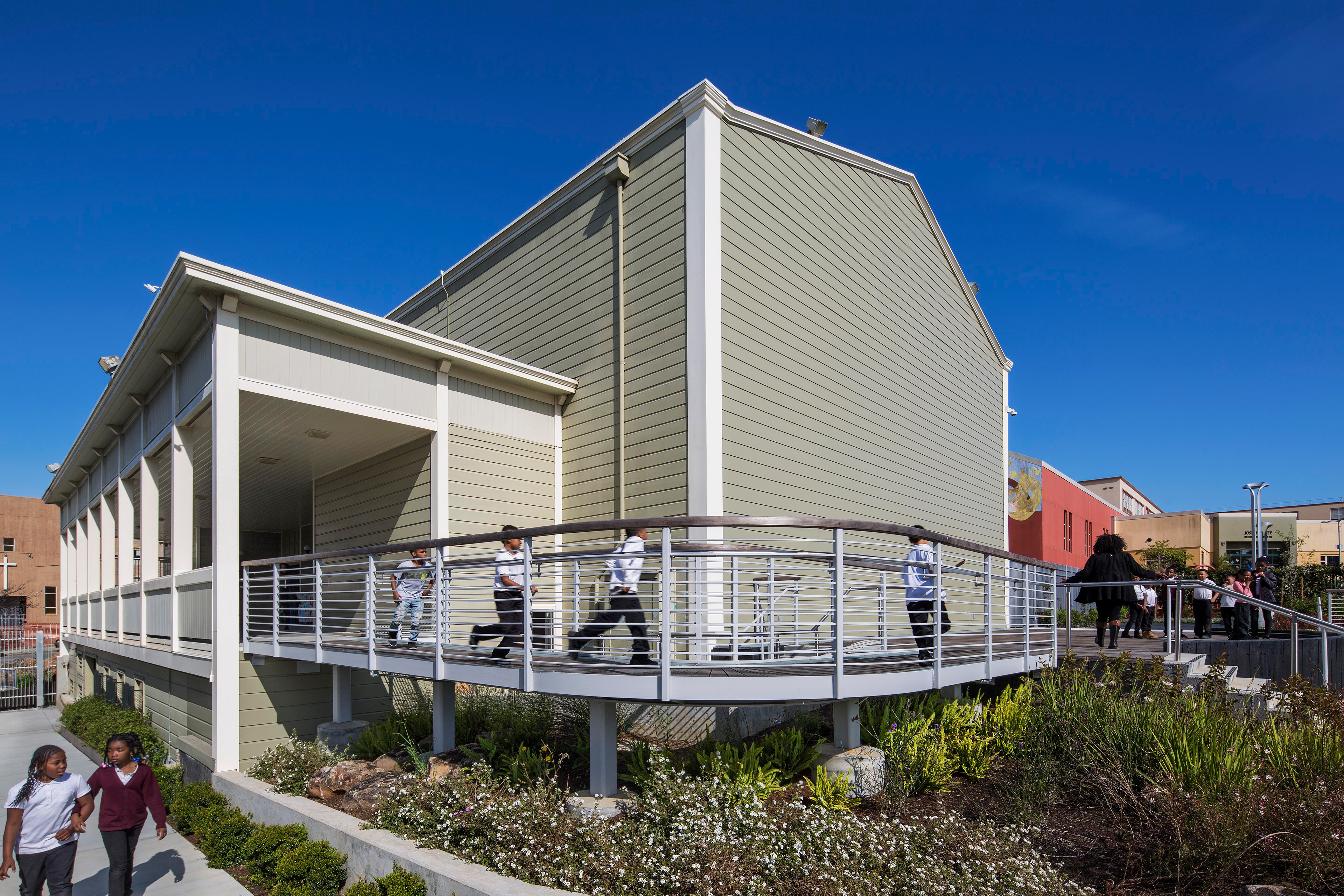 Children run along a curved accessible ramp leading to the South San Francisco Opera House, a restored historic building with sage-green siding and white trim. The ramp is surrounded by landscaped greenery under a bright blue sky, highlighting the structure’s welcoming community design.