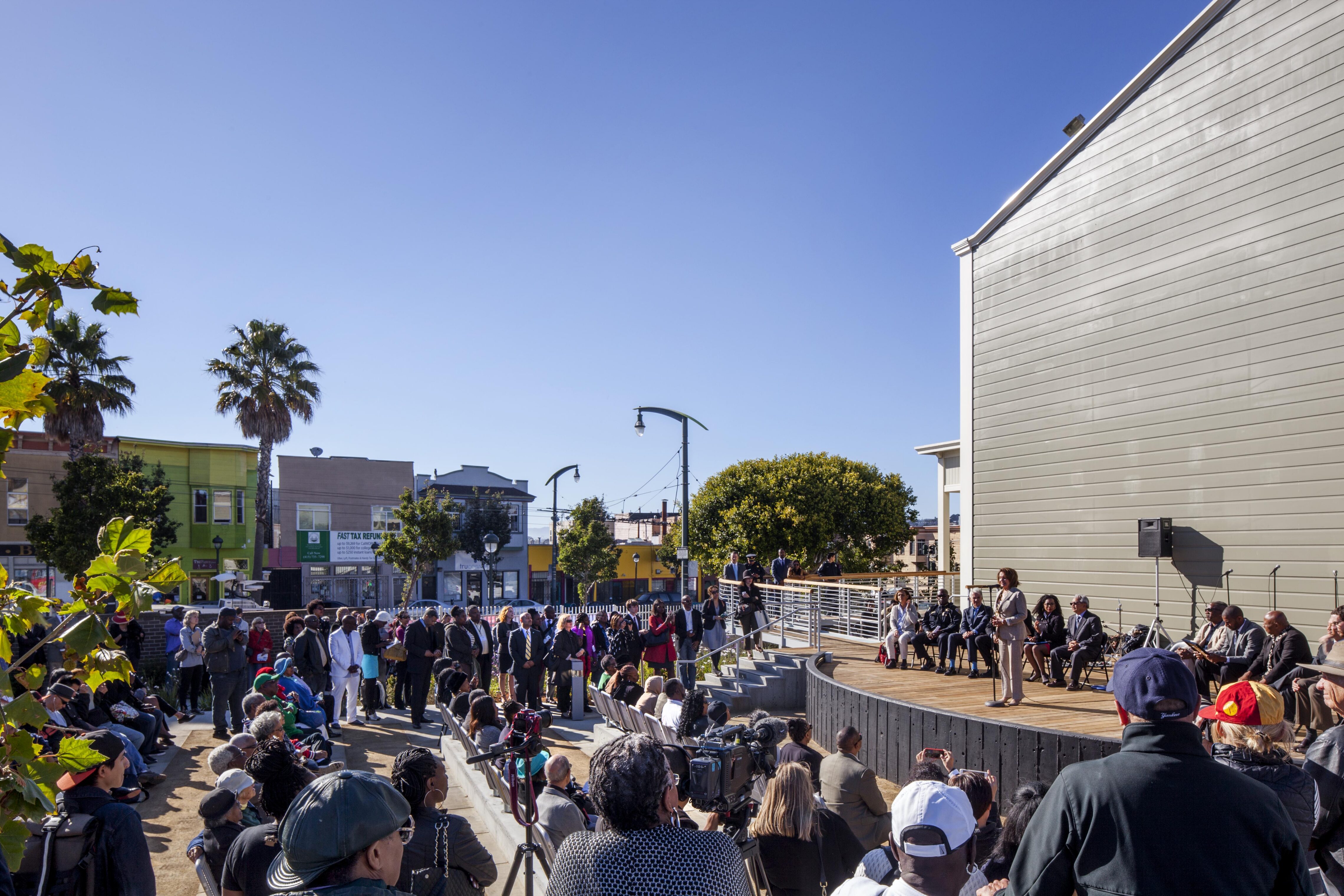A large crowd gathers outdoors for a community event in front of the South San Francisco Opera House. People sit and stand around an open-air stage where a speaker addresses the audience under clear blue skies, surrounded by colorful neighborhood buildings and palm trees.
