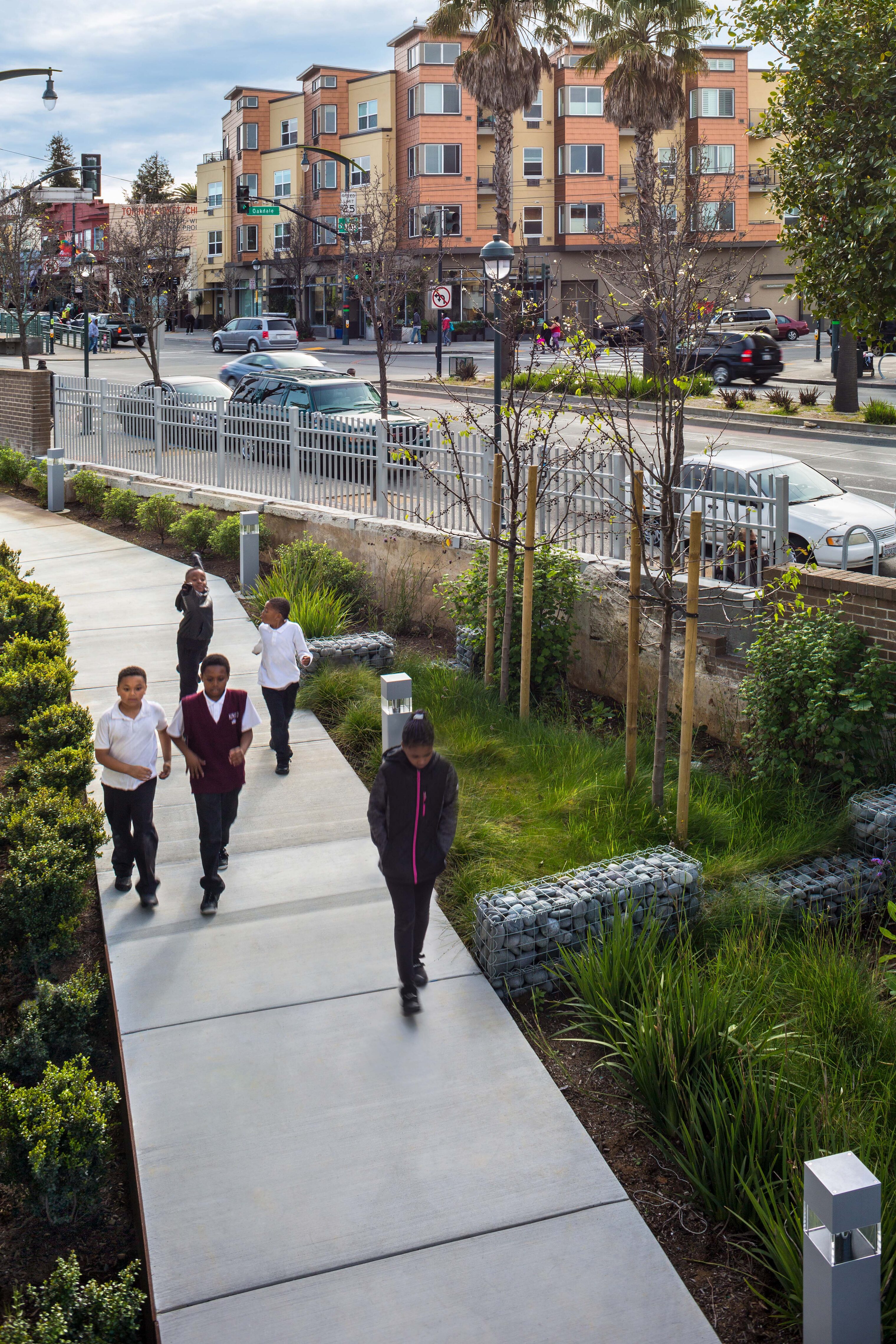 Children walk along a landscaped pathway bordered by greenery, gabion benches, and young trees. The scene overlooks a busy urban street lined with colorful apartment buildings and palm trees, capturing a lively neighborhood setting.