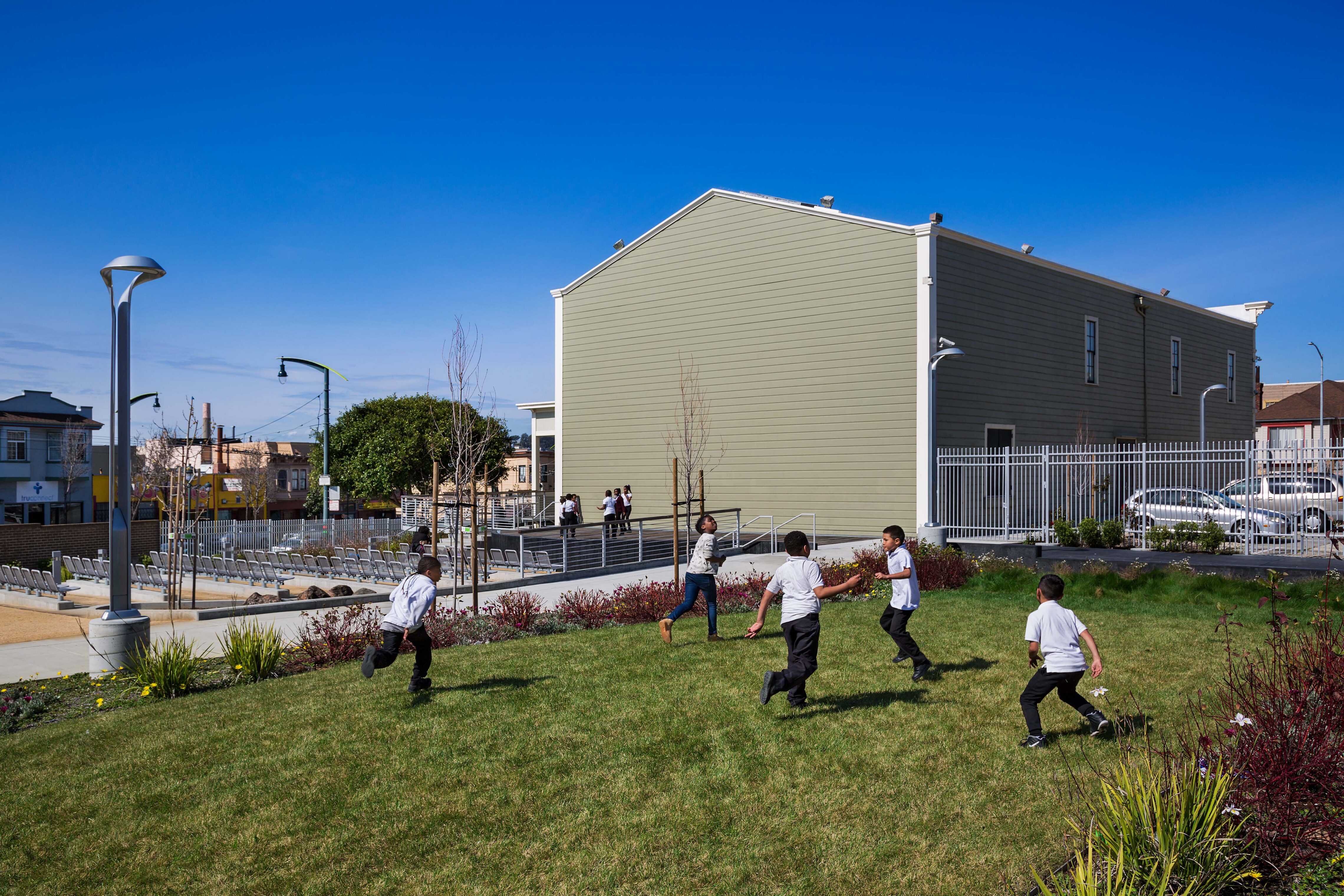 Children play a game of tag on a grassy lawn beside a light green building under a bright blue sky. The landscaped outdoor area features pathways, lighting, and seating in the background, creating an inviting and active community environment.