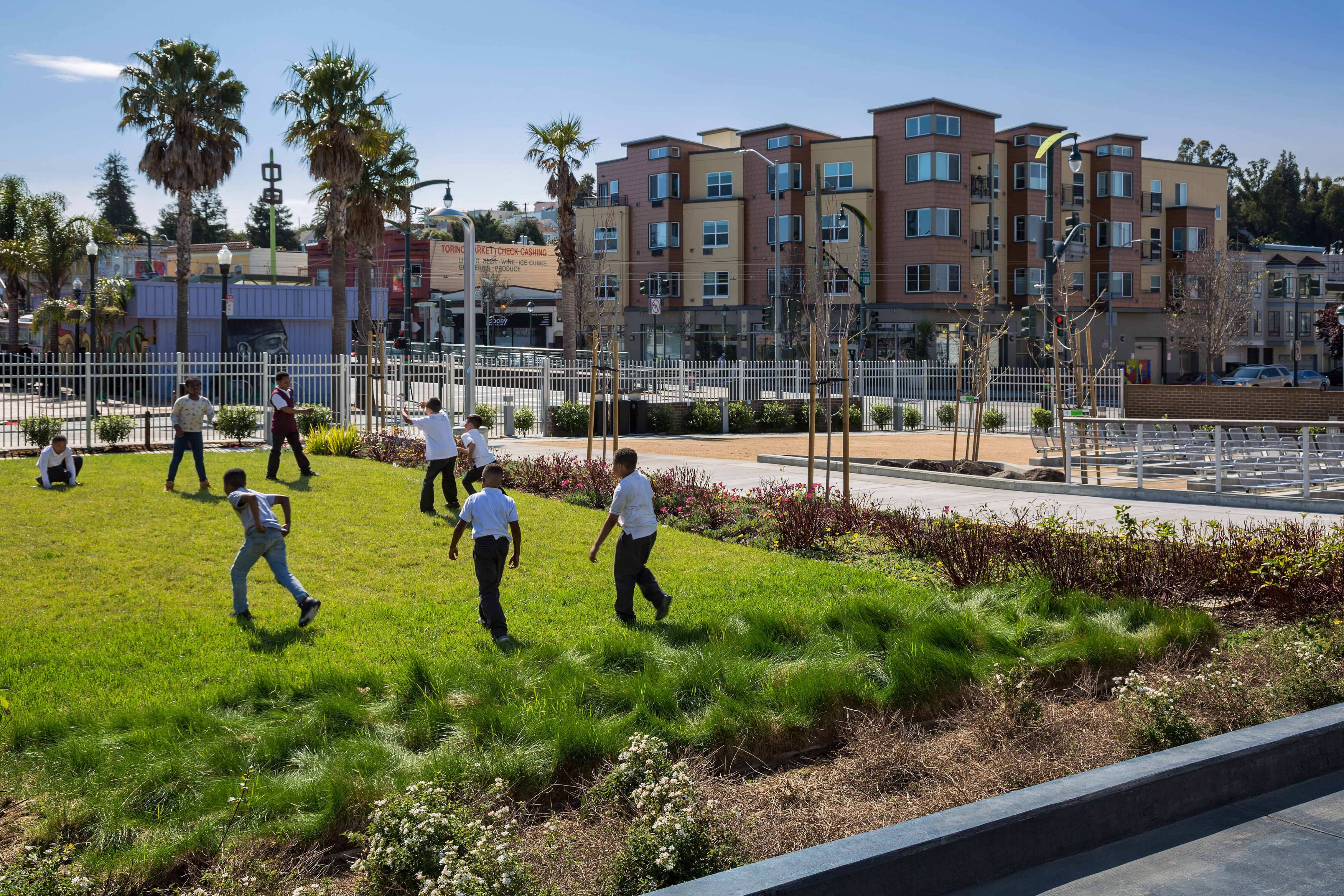 Children play energetically on a sunny grassy lawn bordered by new landscaping and young trees. In the background, palm trees, colorful urban buildings, and street activity create a lively neighborhood atmosphere.