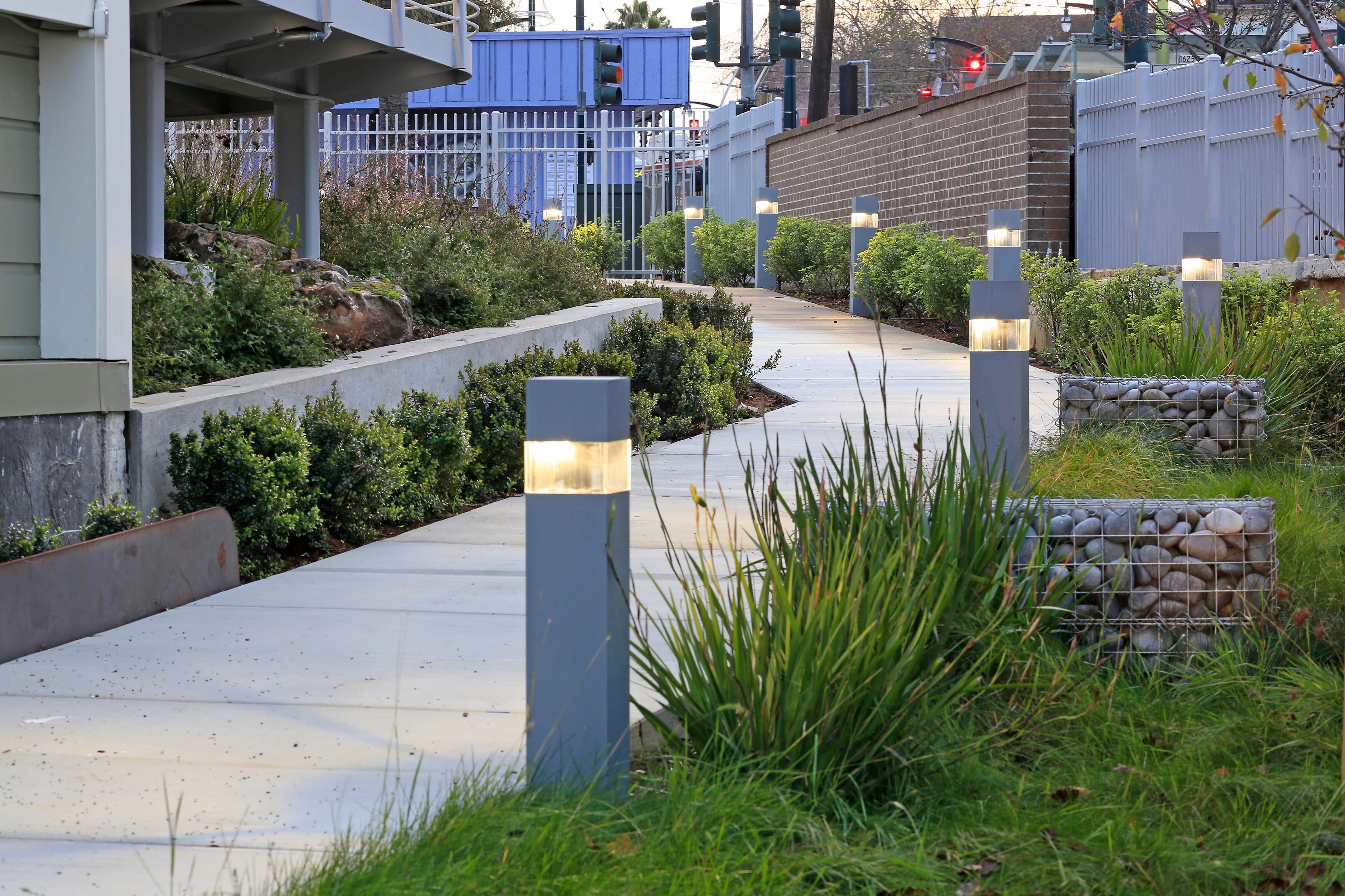 Concrete walkway lined with illuminated bollard lights, low greenery, and gabion stone planters next to a building and brick wall.
