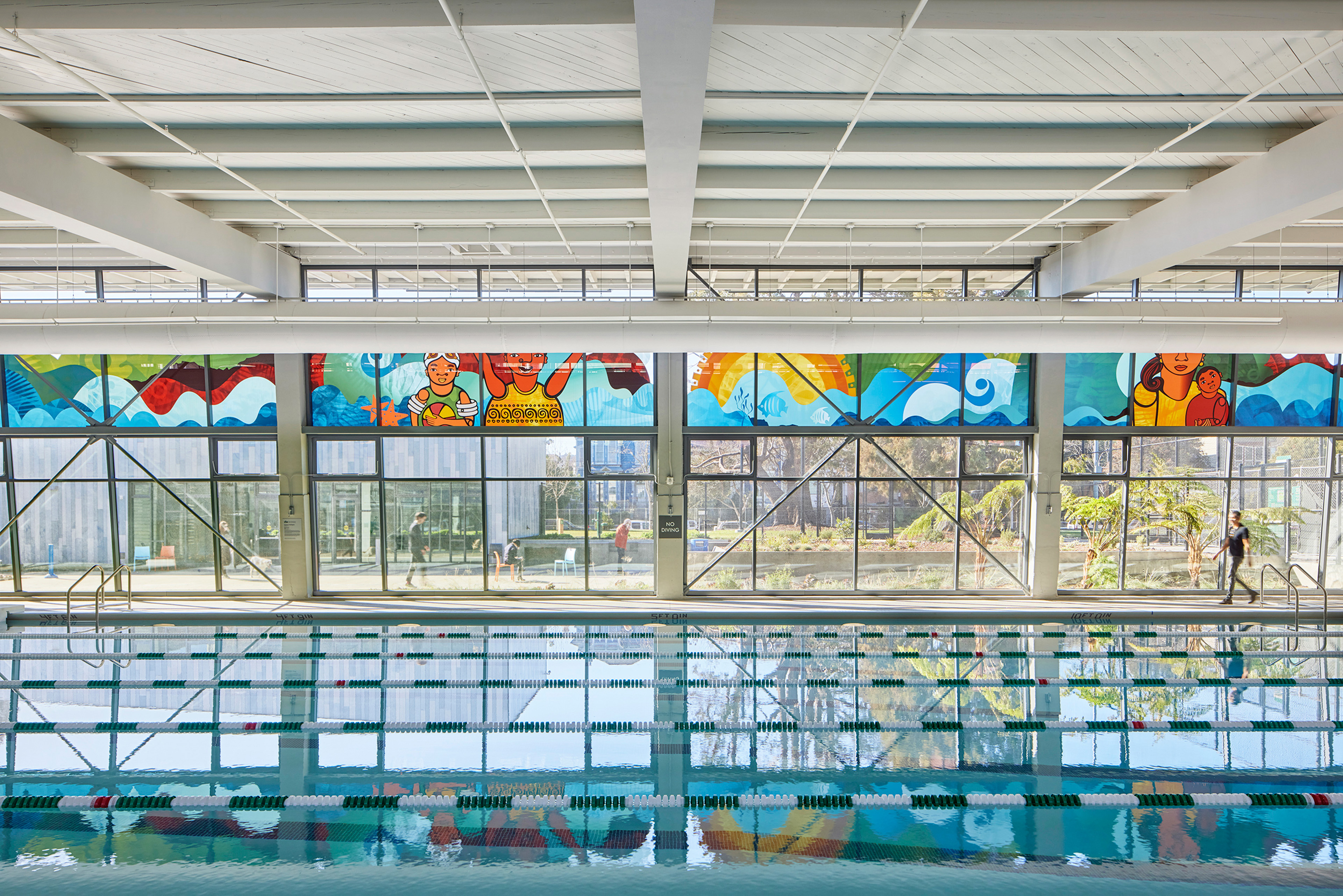 Indoor swimming pool with green-and-white lane dividers, surrounded by large windows and colorful mural panels depicting swimmers and ocean waves. Sunlight streams through the glass, reflecting vibrant colors on the water’s surface.