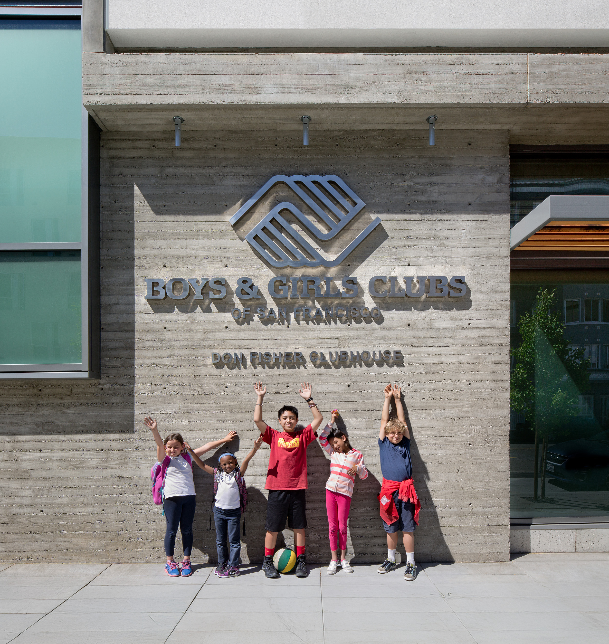Five children stand smiling with their hands raised in front of the concrete exterior wall of the Boys & Girls Clubs of San Francisco’s Don Fisher Clubhouse. The organization’s logo and name are prominently displayed above them in metal lettering.